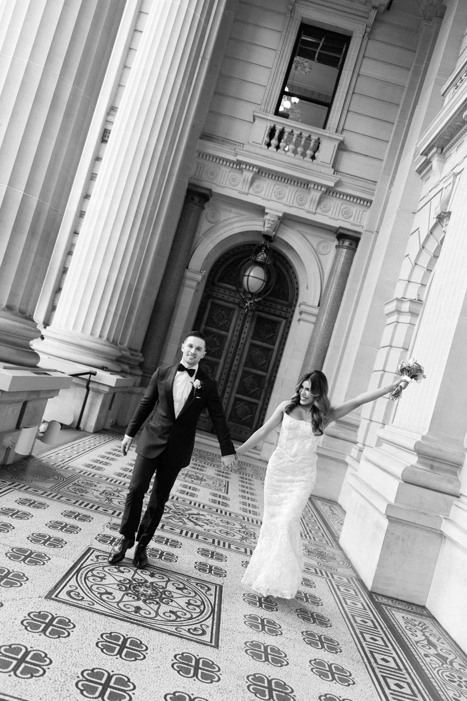 Black and white photo of a bride and groom holding hands outside a grand classical building with ornate columns.