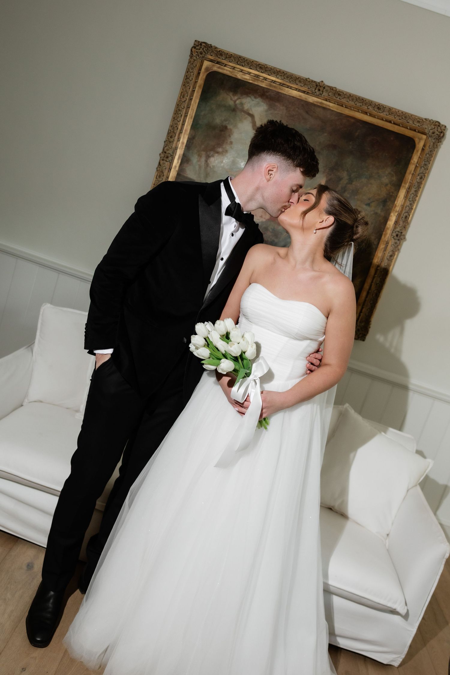 Bride and groom sharing a kiss in elegant wedding attire, bride holding white tulip bouquet before a classic painting.