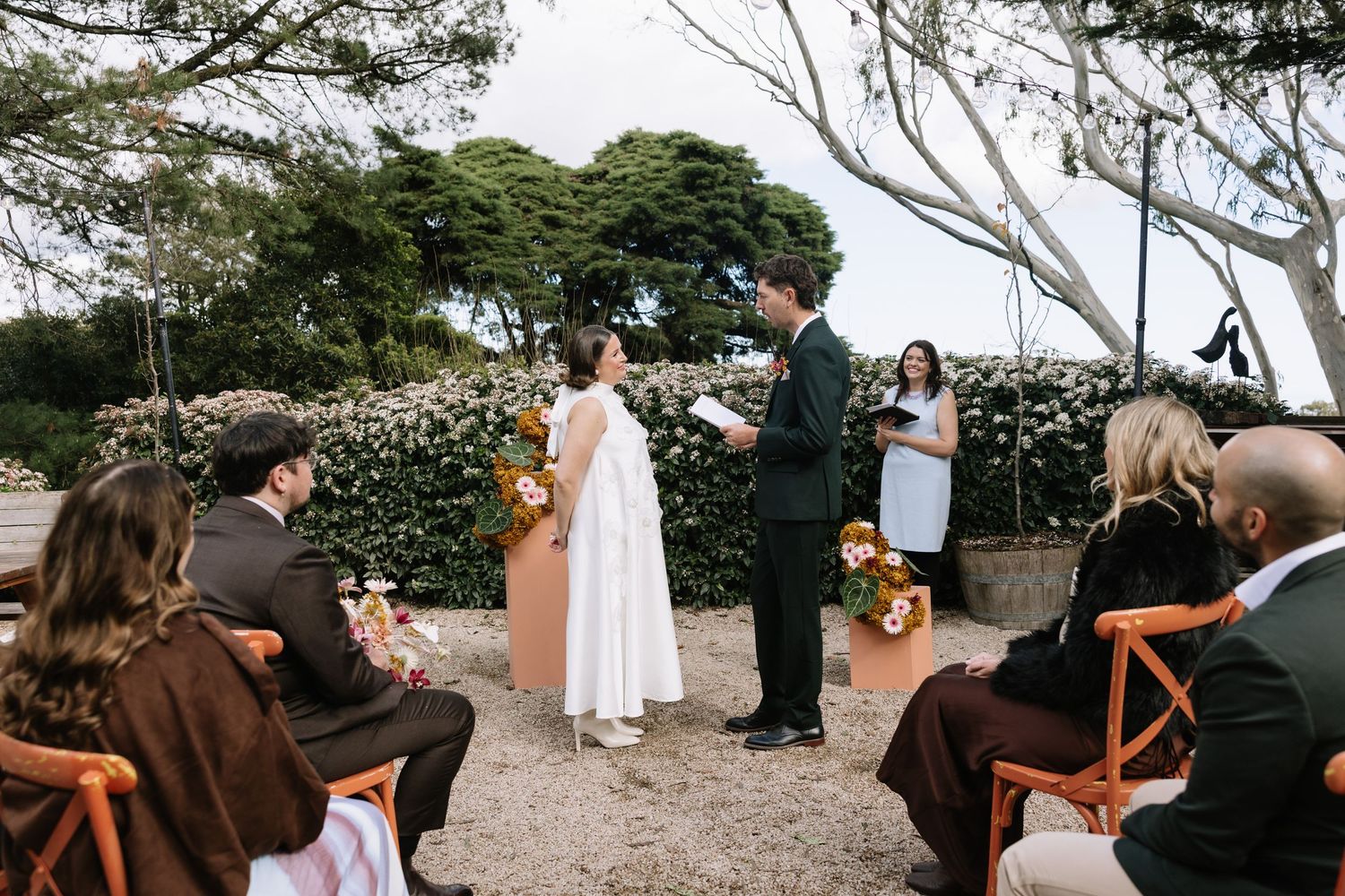 Outdoor wedding ceremony with couple exchanging vows before officiant, guests seated on wooden chairs amid lush greenery.