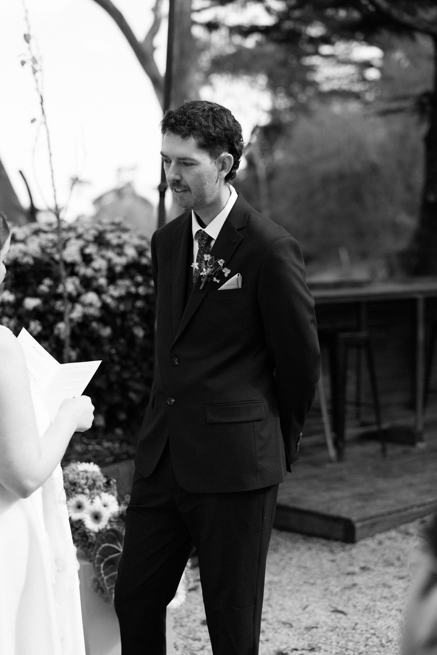Black and white photo of a groom in a suit with boutonniere smiling during an outdoor wedding ceremony.