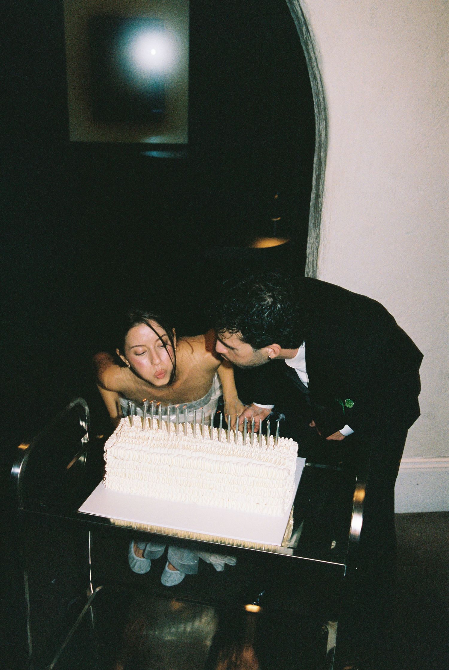 Bride and groom leaning in to blow out candles on white wedding cake at evening reception.
