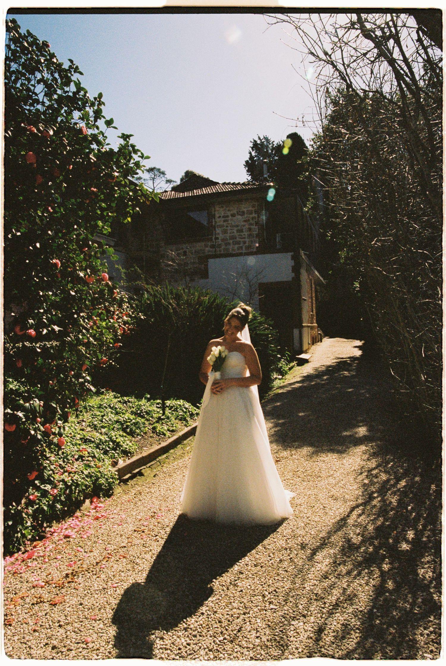 Bride in flowing white gown walks along a sunlit garden path surrounded by colorful blooms and greenery.