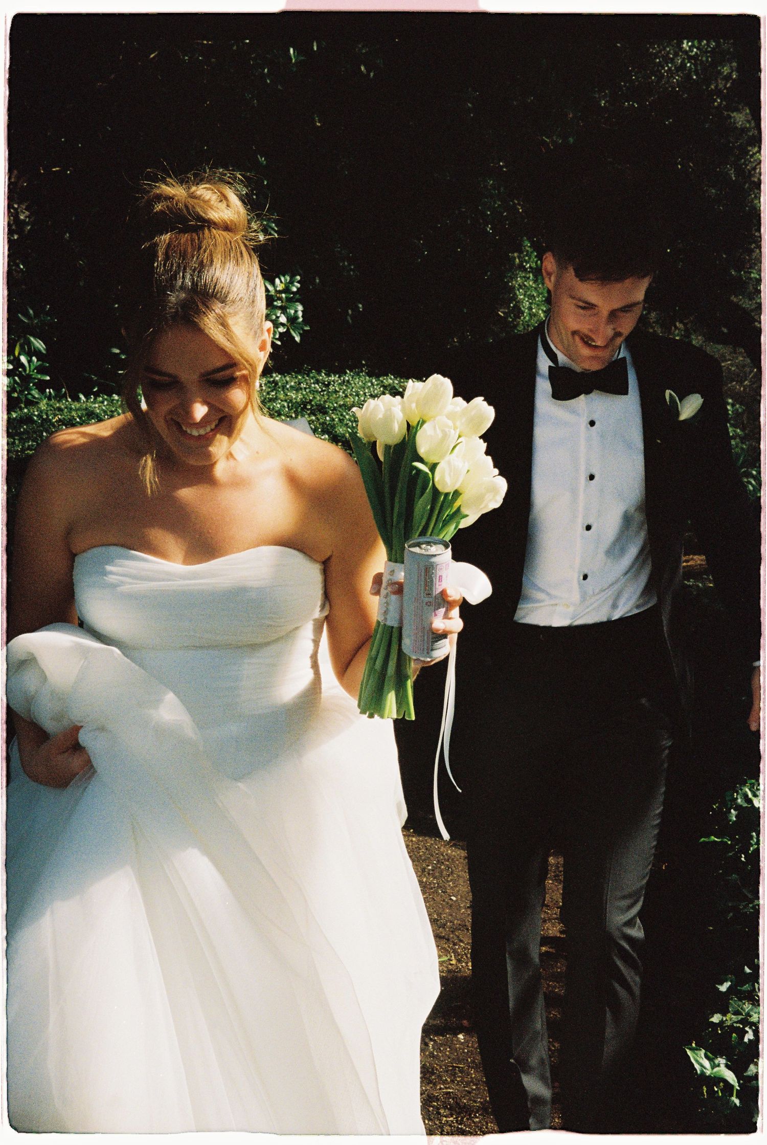 Joyful bride in strapless white gown and groom carry white tulip bouquet along garden pathway.