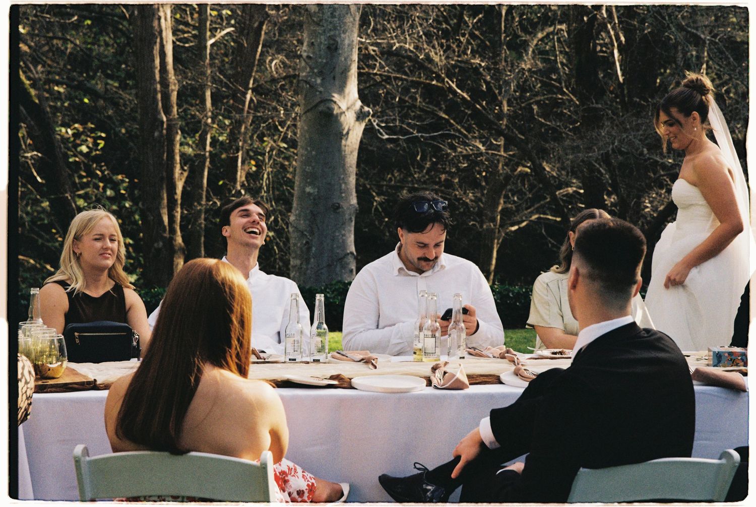 Wedding guests laugh and smile at outdoor garden reception table with bride in white dress nearby.
