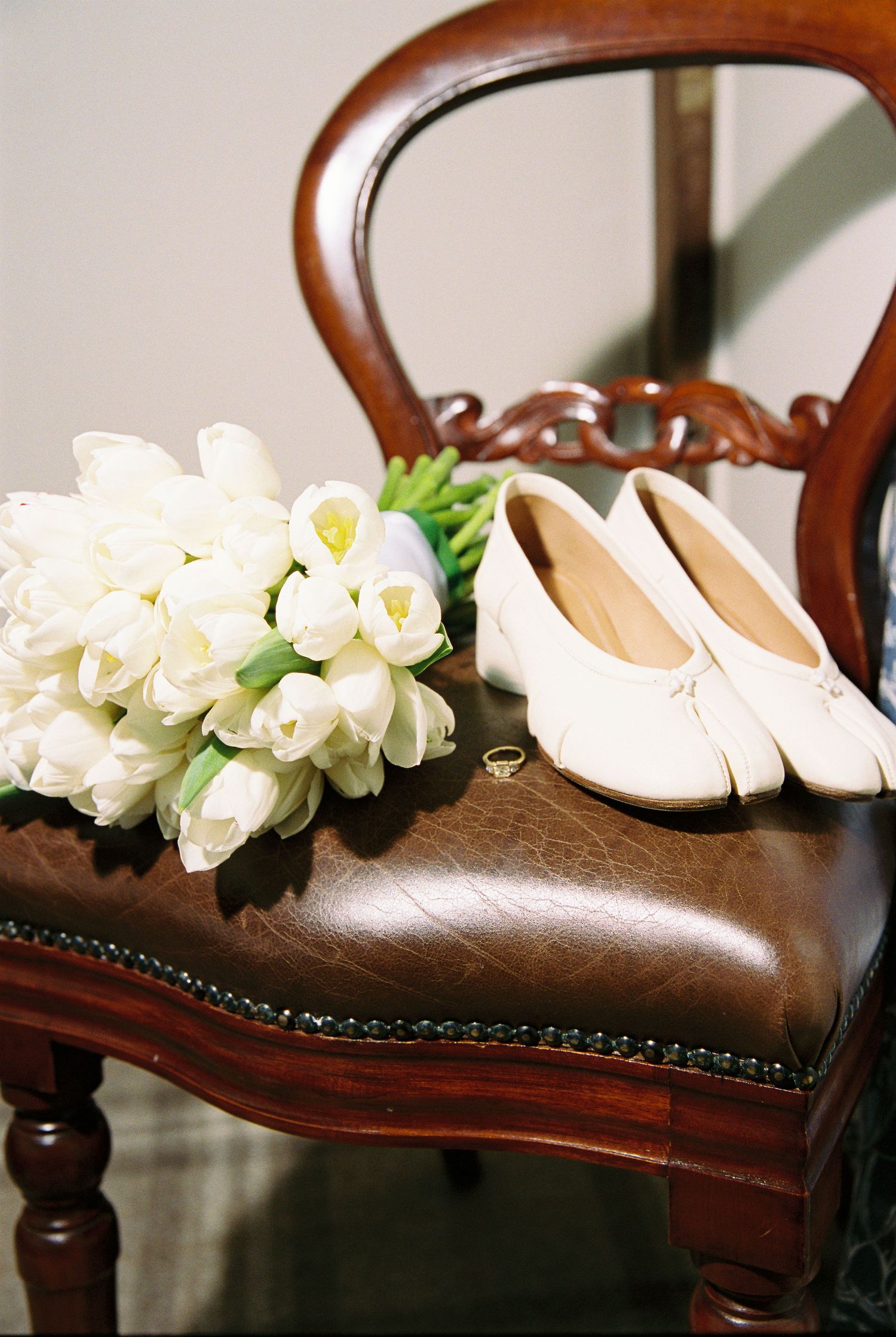 White tulip bridal bouquet, gold ring, and ivory flat bridal shoes arranged on a leather wooden chair.