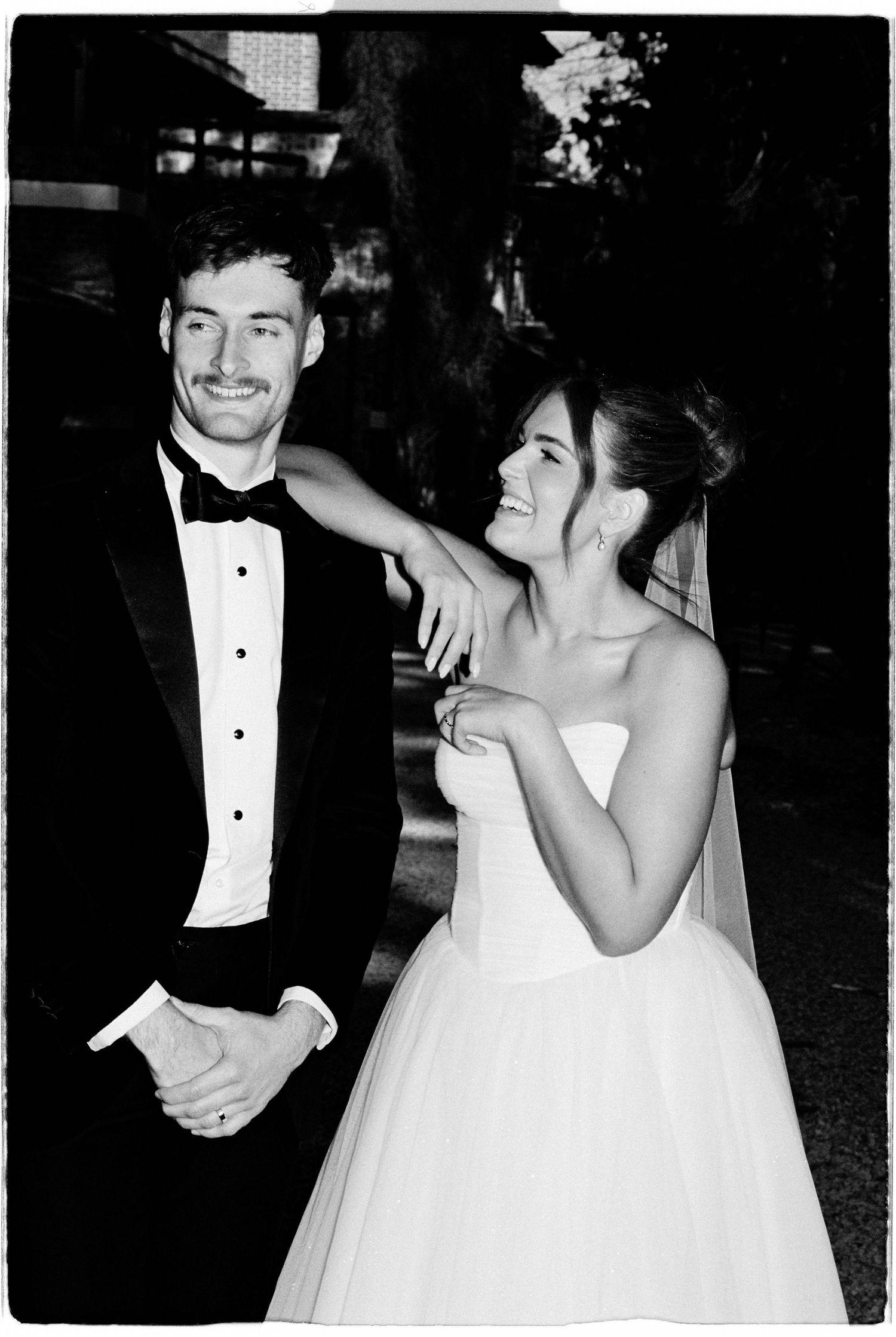 Black and white photo of smiling bride and groom in tuxedo posing together outdoors at night.