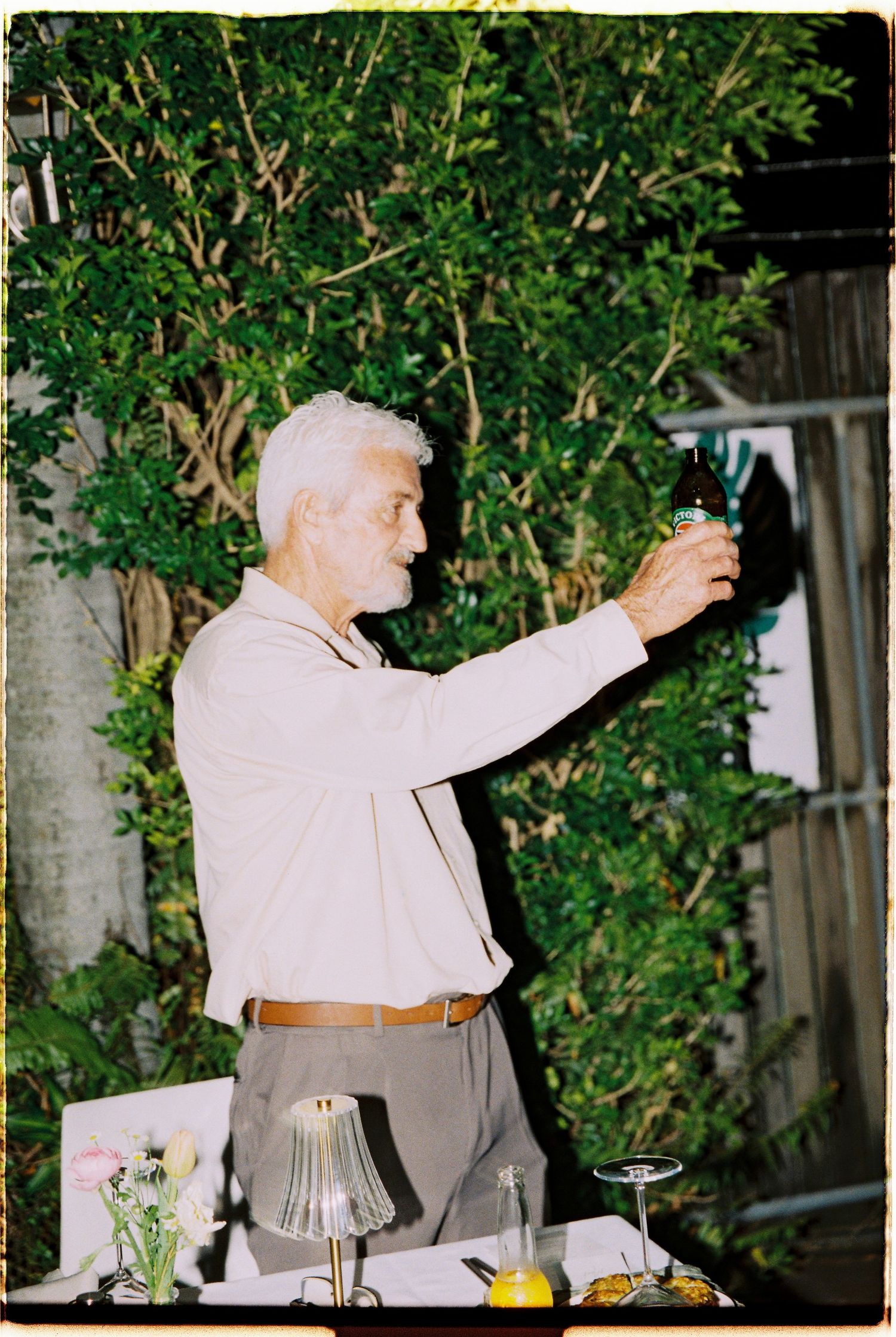 Silver-haired man in beige shirt raising beer bottle toast at outdoor wedding reception with greenery backdrop.
