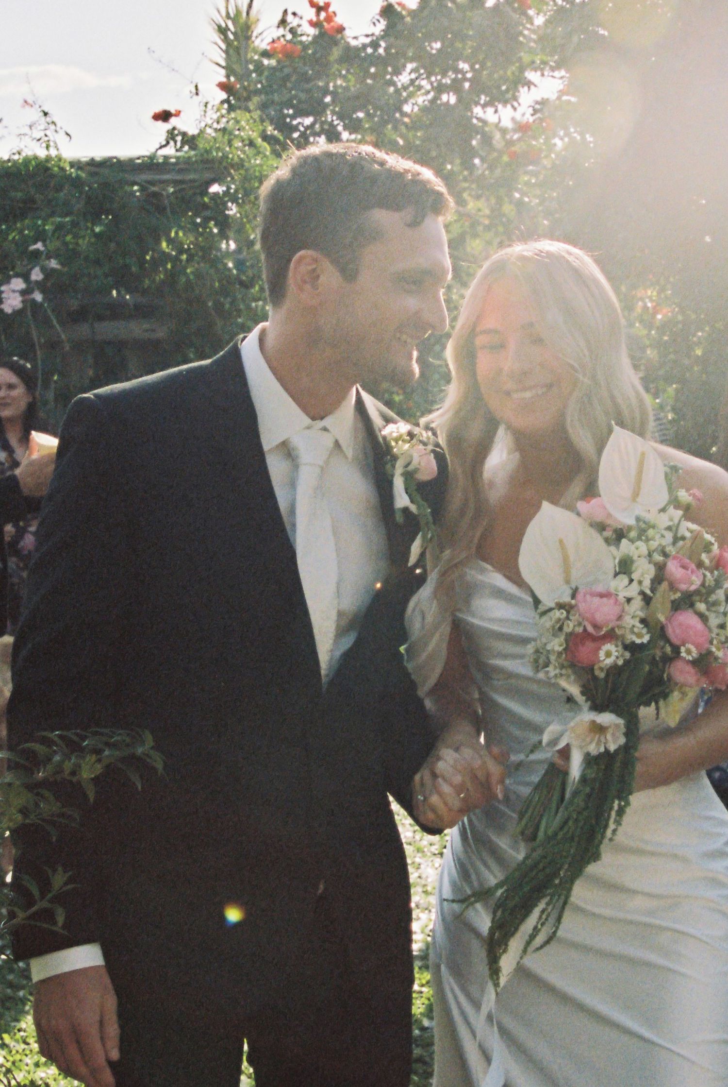 Smiling newlyweds walk together in golden sunlight after outdoor garden ceremony.