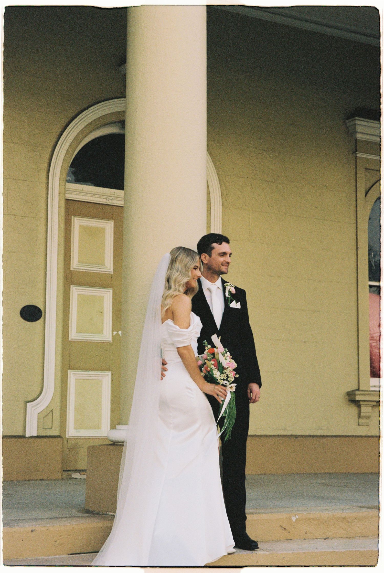 Bride and groom pose on church steps, bride in off-shoulder white gown with veil, groom in black tuxedo.