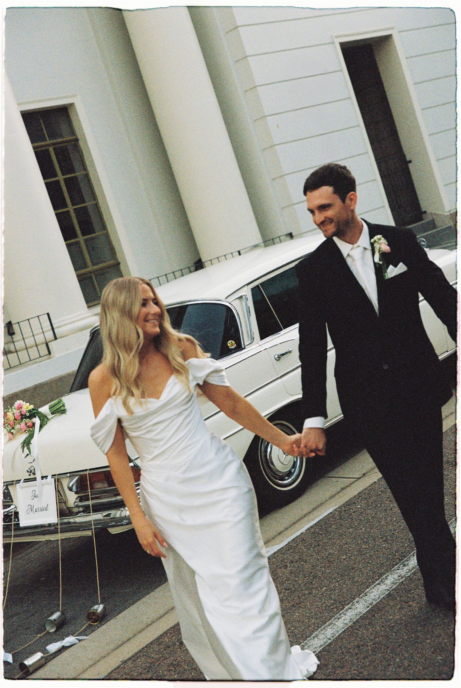 Smiling bride and groom holding hands walking beside a classic white vintage wedding car outside a grand venue.