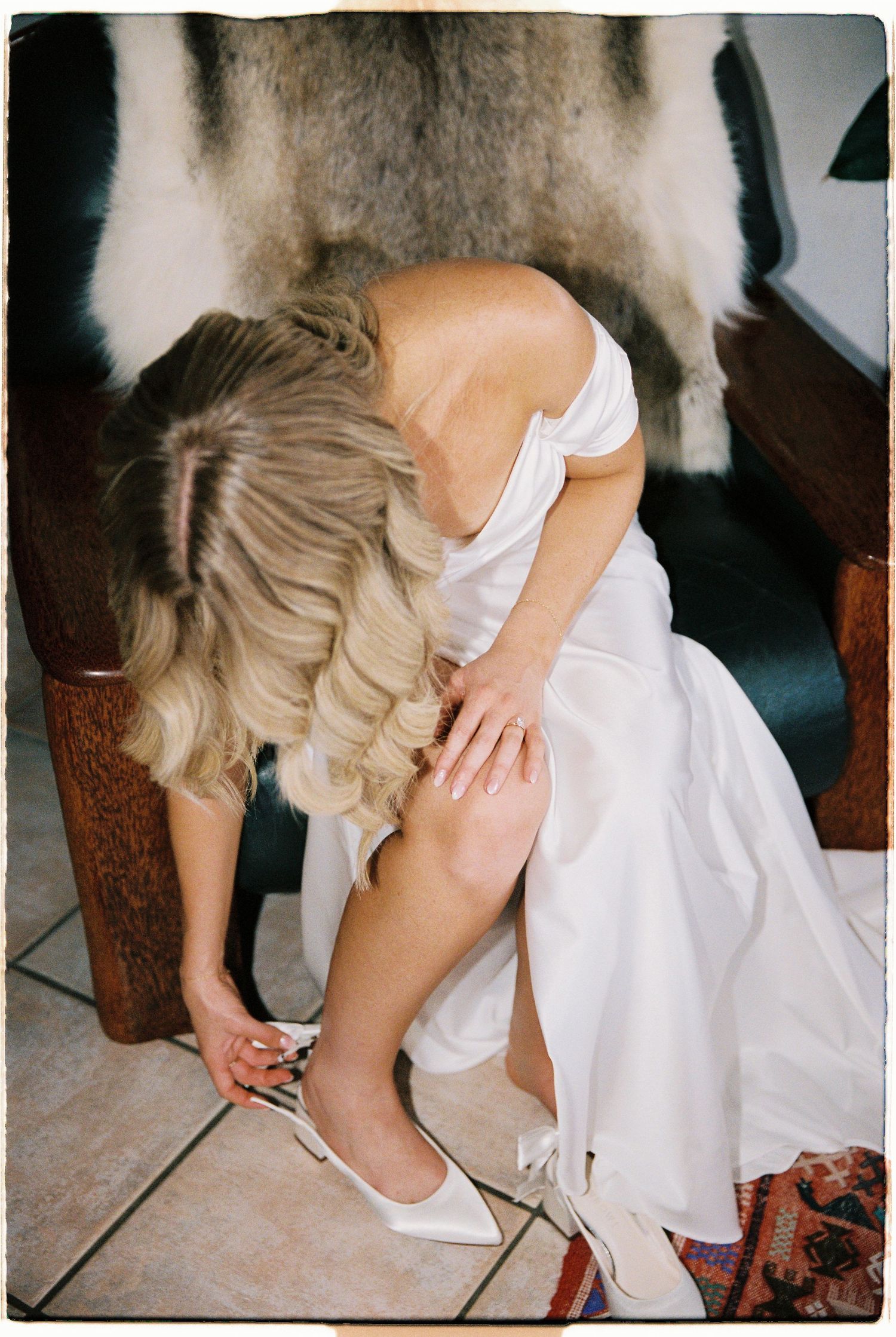 Bride in white off-shoulder gown putting on white heels while seated on leather chair.
