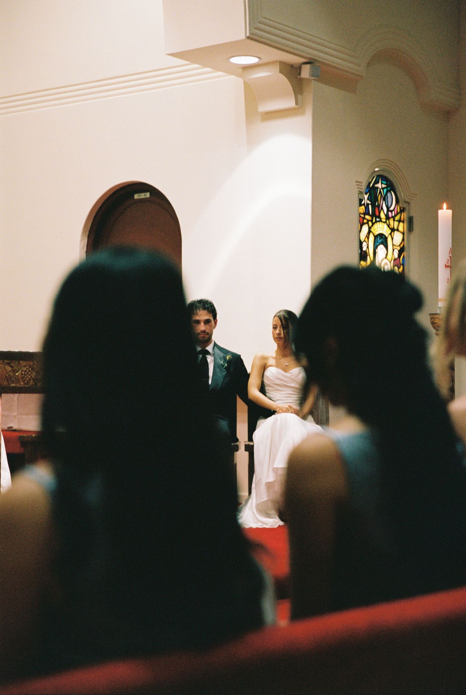 Bride and groom seated at church altar during wedding ceremony with stained glass window in background.