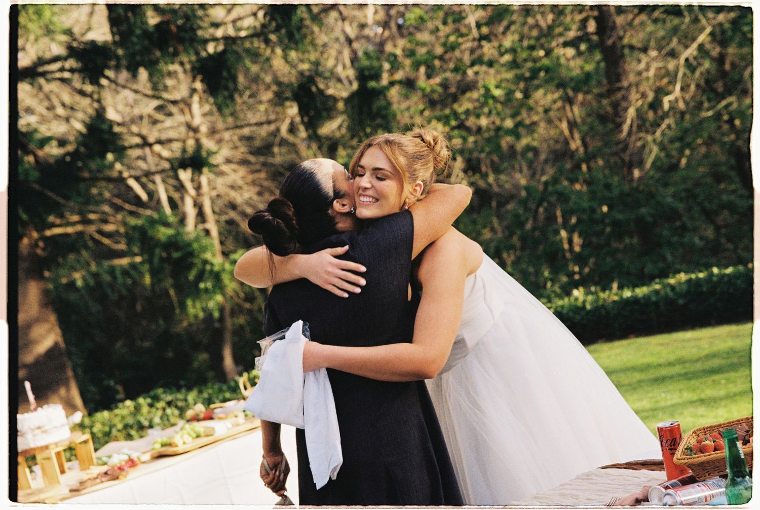 Bride in white gown hugging a guest in black dress at an outdoor wedding reception.