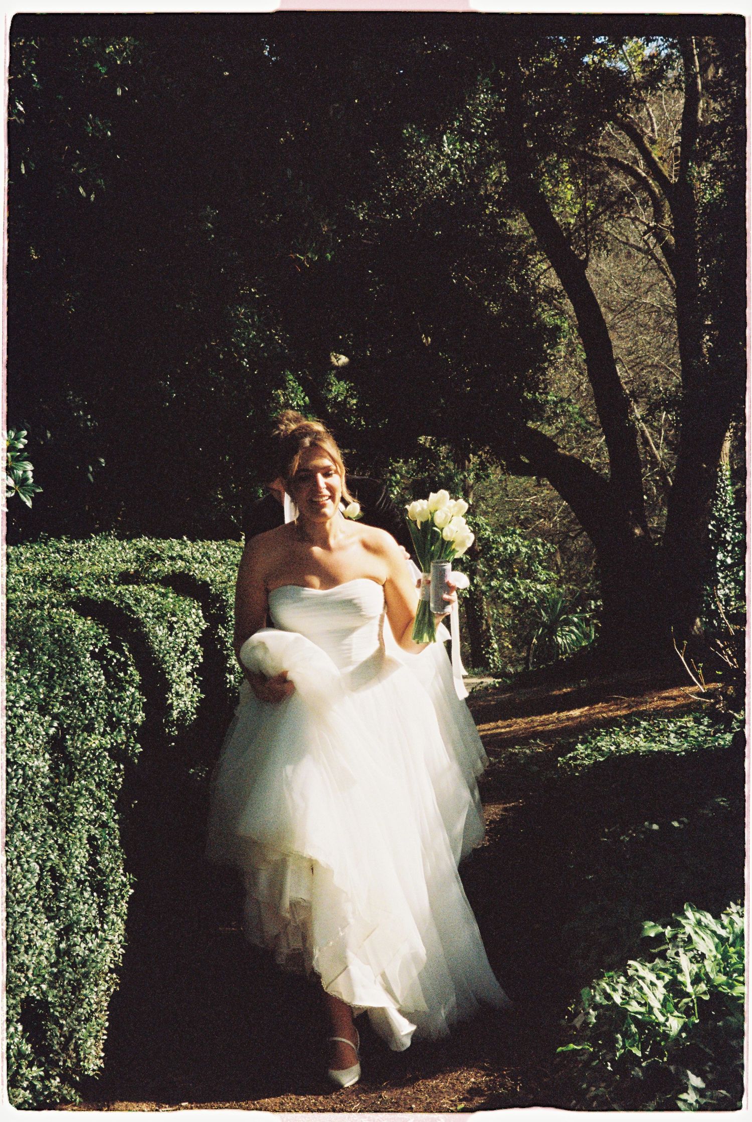 A bride in a white tulle gown walks through a garden path holding a bouquet.