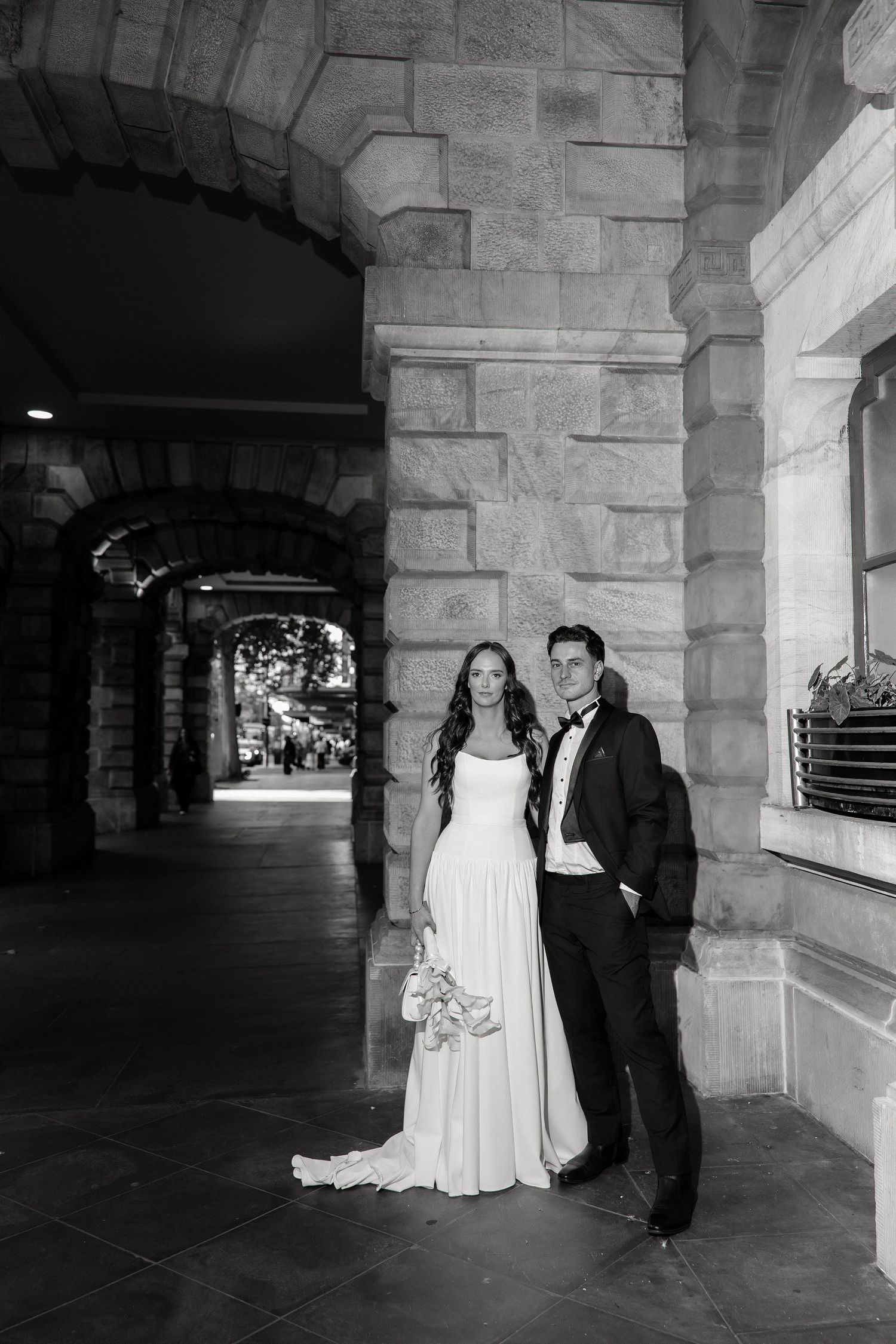 Bride and groom stand elegantly under stone arches of a heritage building in a black and white wedding photo.