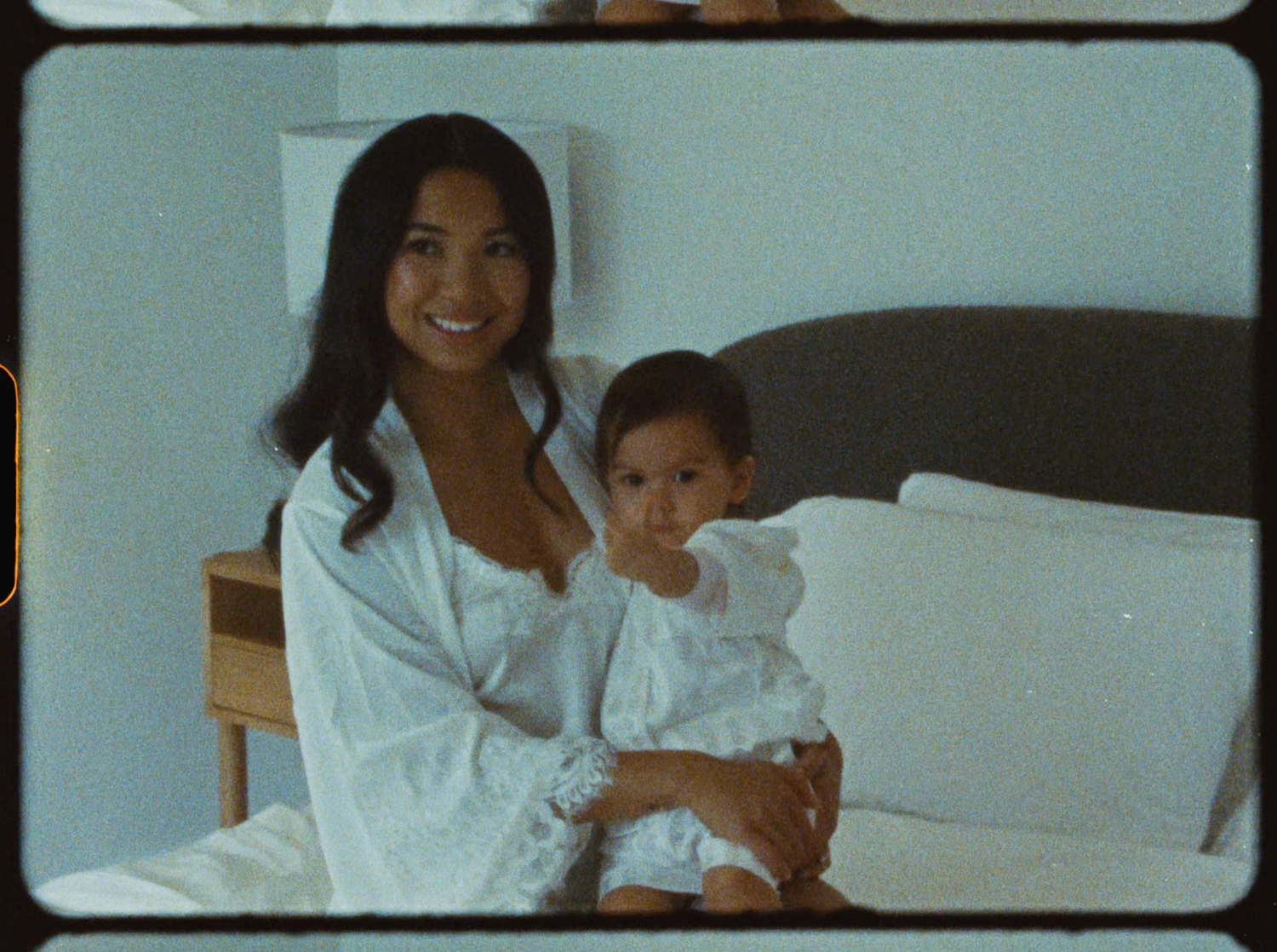 Smiling woman in white robe sitting on bed holding a baby dressed in white outfit.