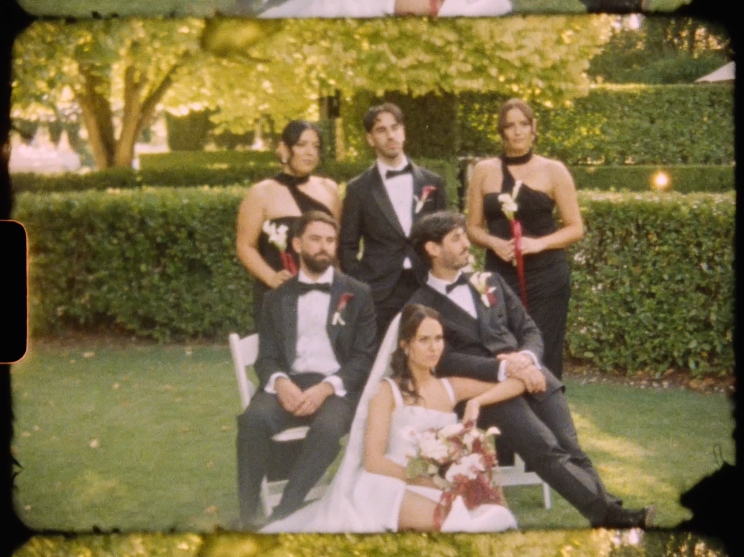 Wedding party portrait featuring bride, groom, bridesmaids in black gowns and groomsmen on garden lawn.