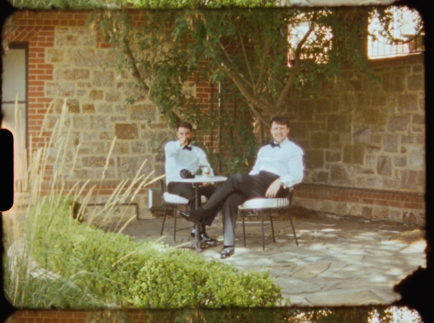 Two men in tuxedos sitting at outdoor patio table against stone wall at wedding venue.