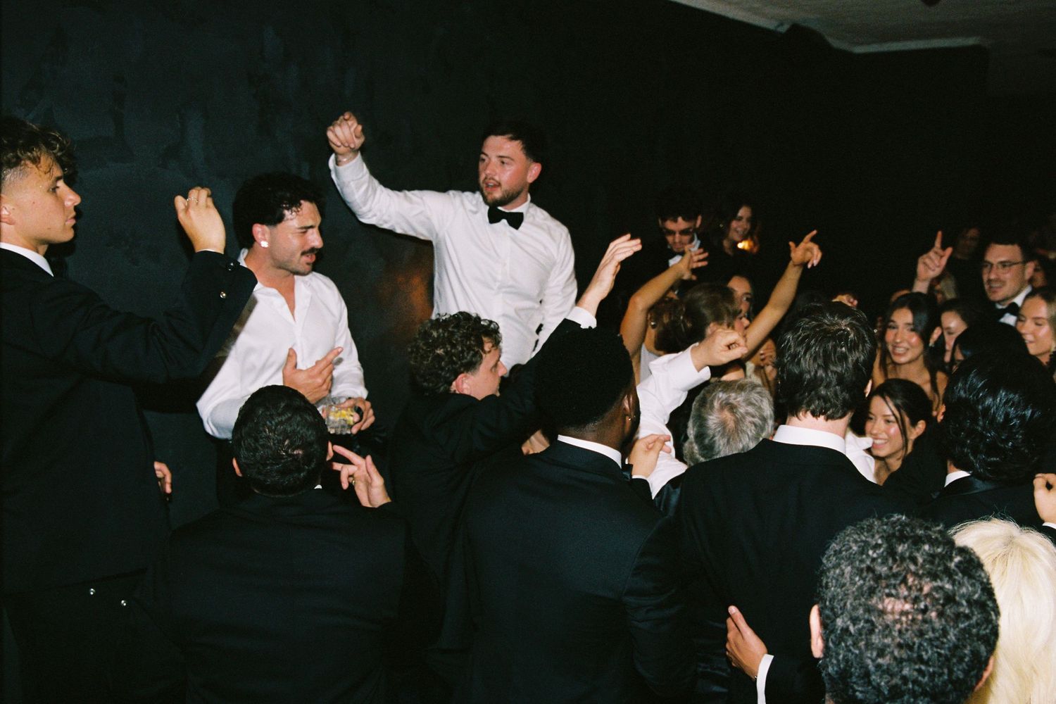 Groom in white shirt and bow tie lifted by cheering guests during lively wedding reception dancing.