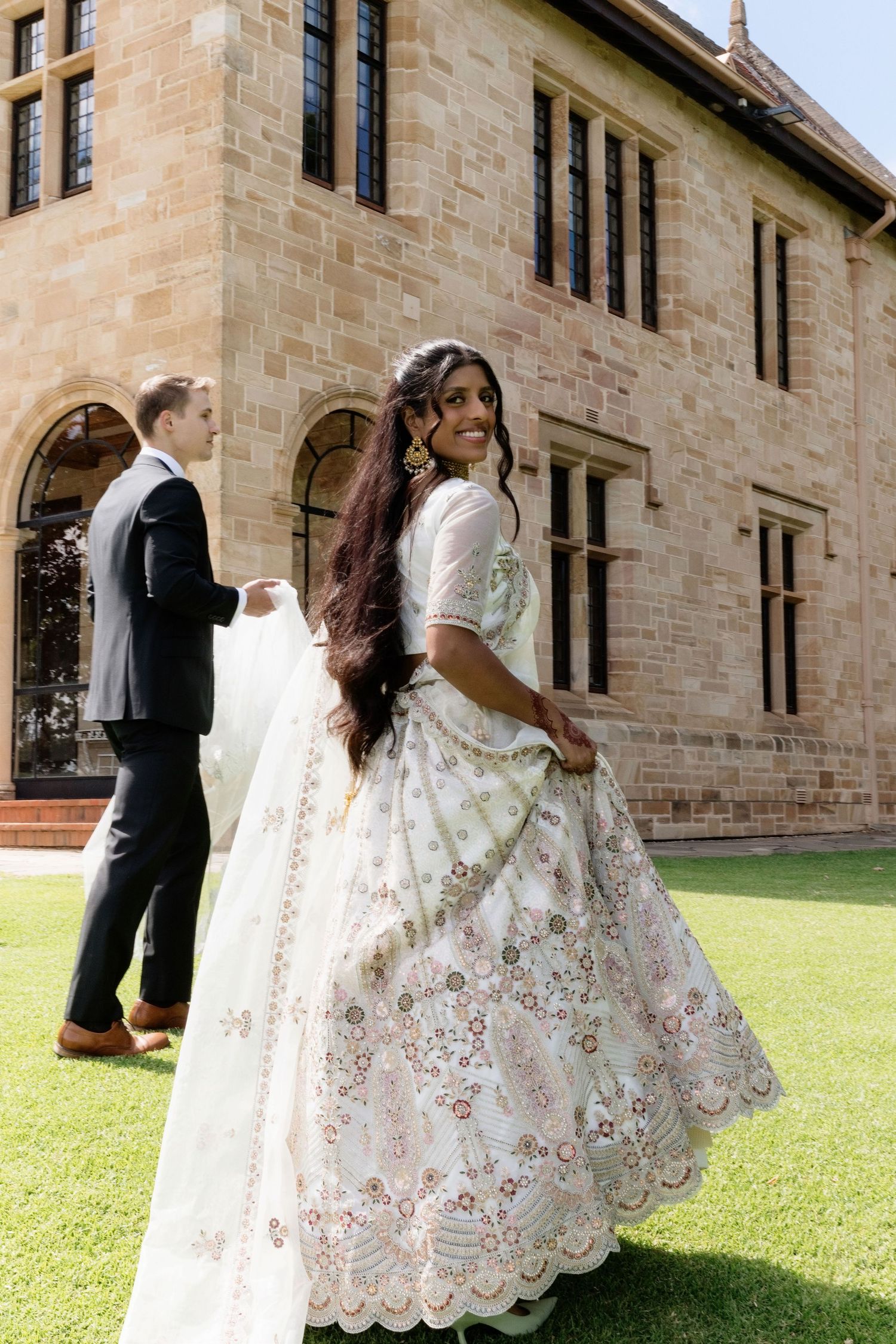 Bride twirling in stunning white embroidered Indian lehenga outside historic sandstone building on her wedding day.