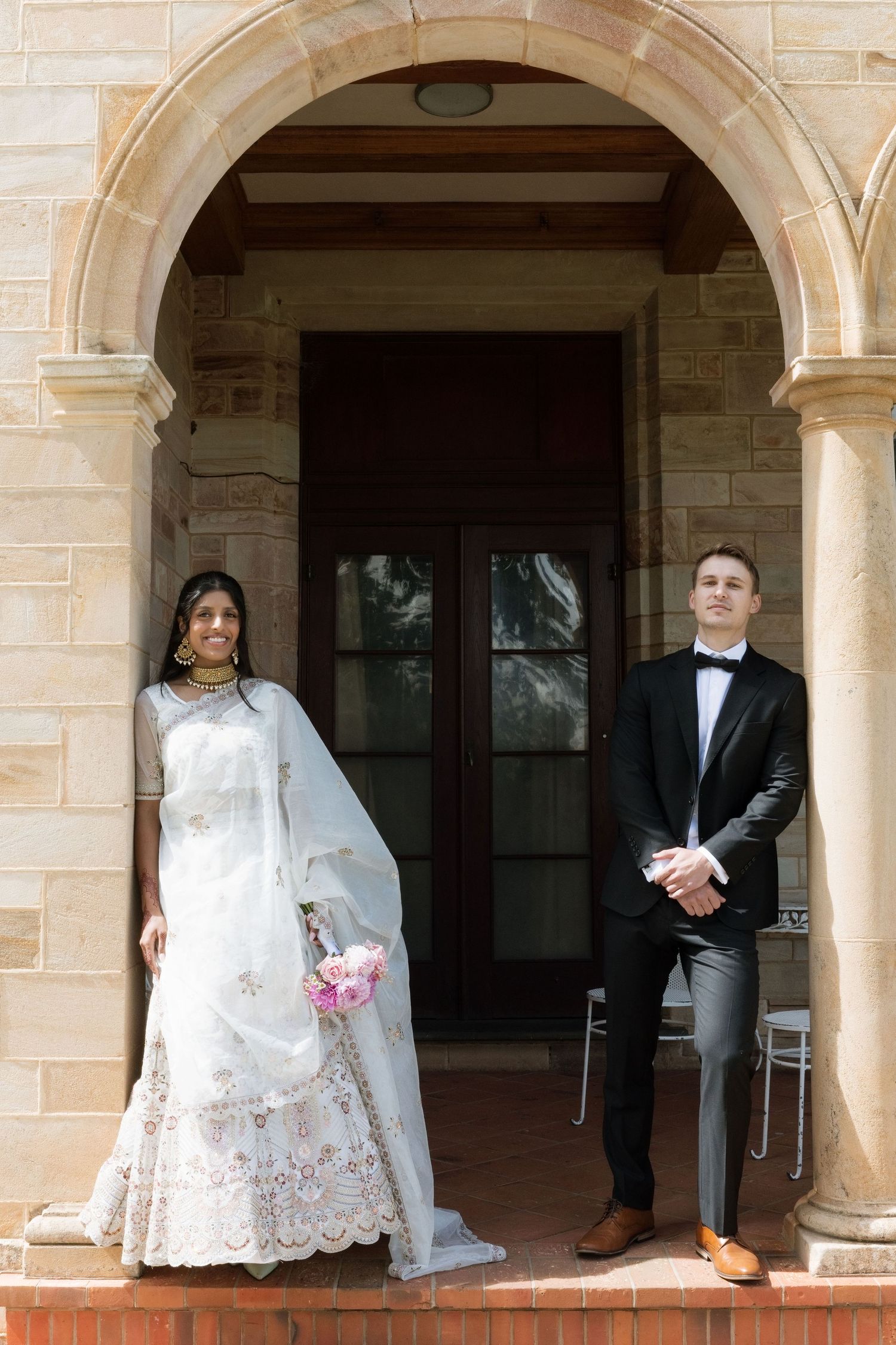 Bride in white Indian lehenga and groom in tuxedo standing under stone archway.