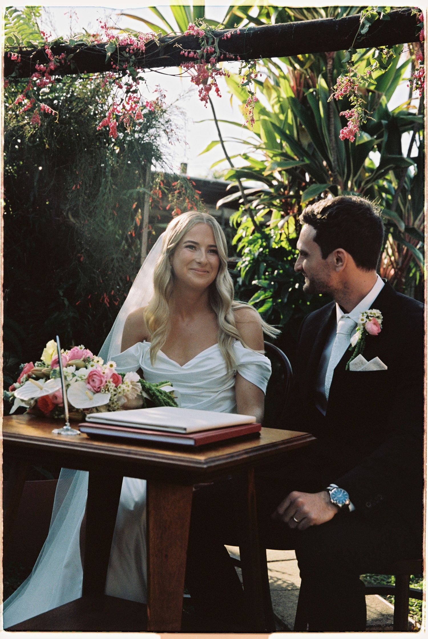 Bride and groom signing wedding register at outdoor ceremony surrounded by tropical greenery and pink flowers.