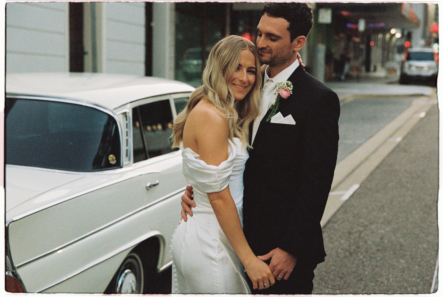 Bride in off-shoulder white gown and groom in black suit pose beside vintage white car on city street.