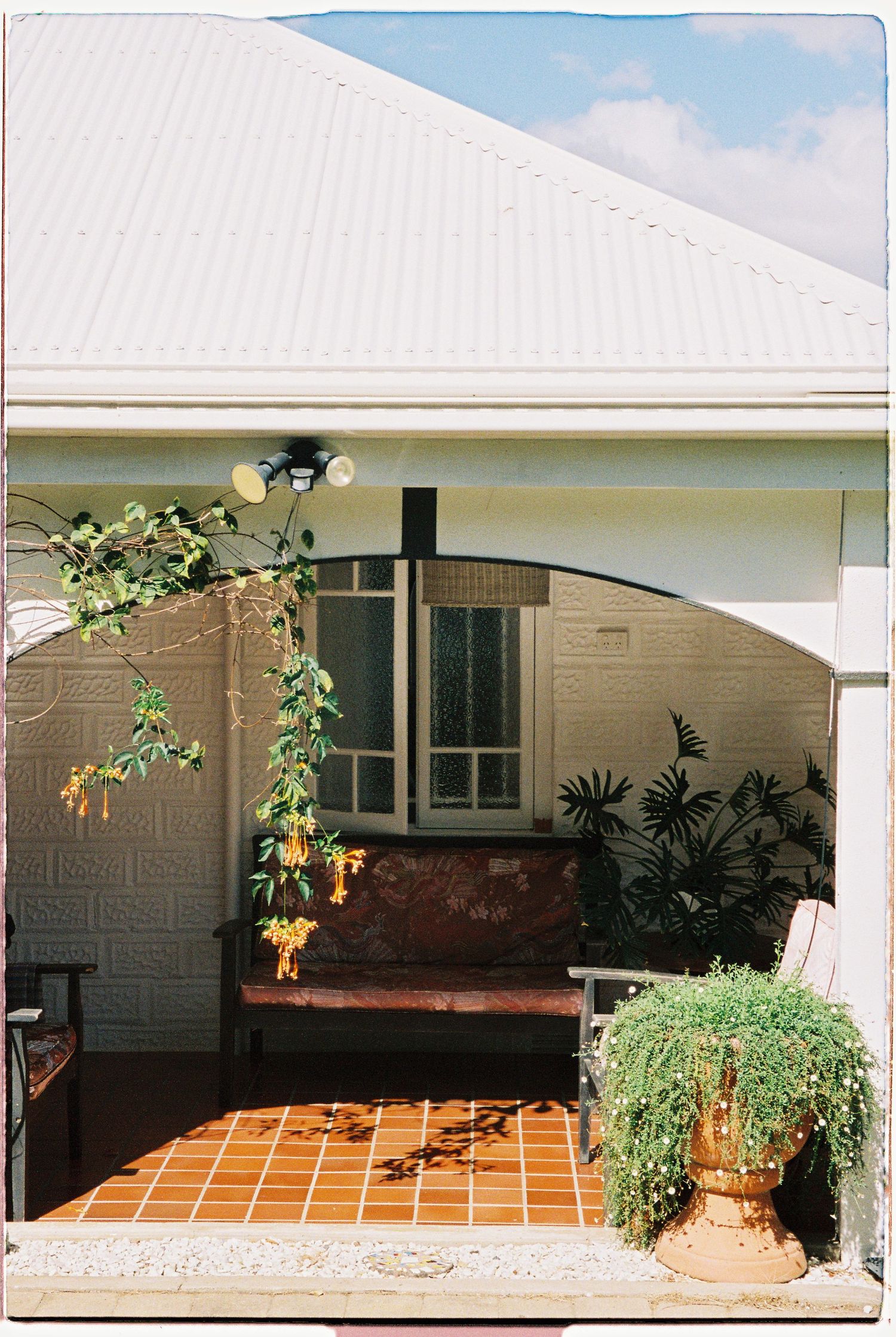 Charming home exterior featuring a covered porch with terracotta tiles, lush greenery, and arched entryway.