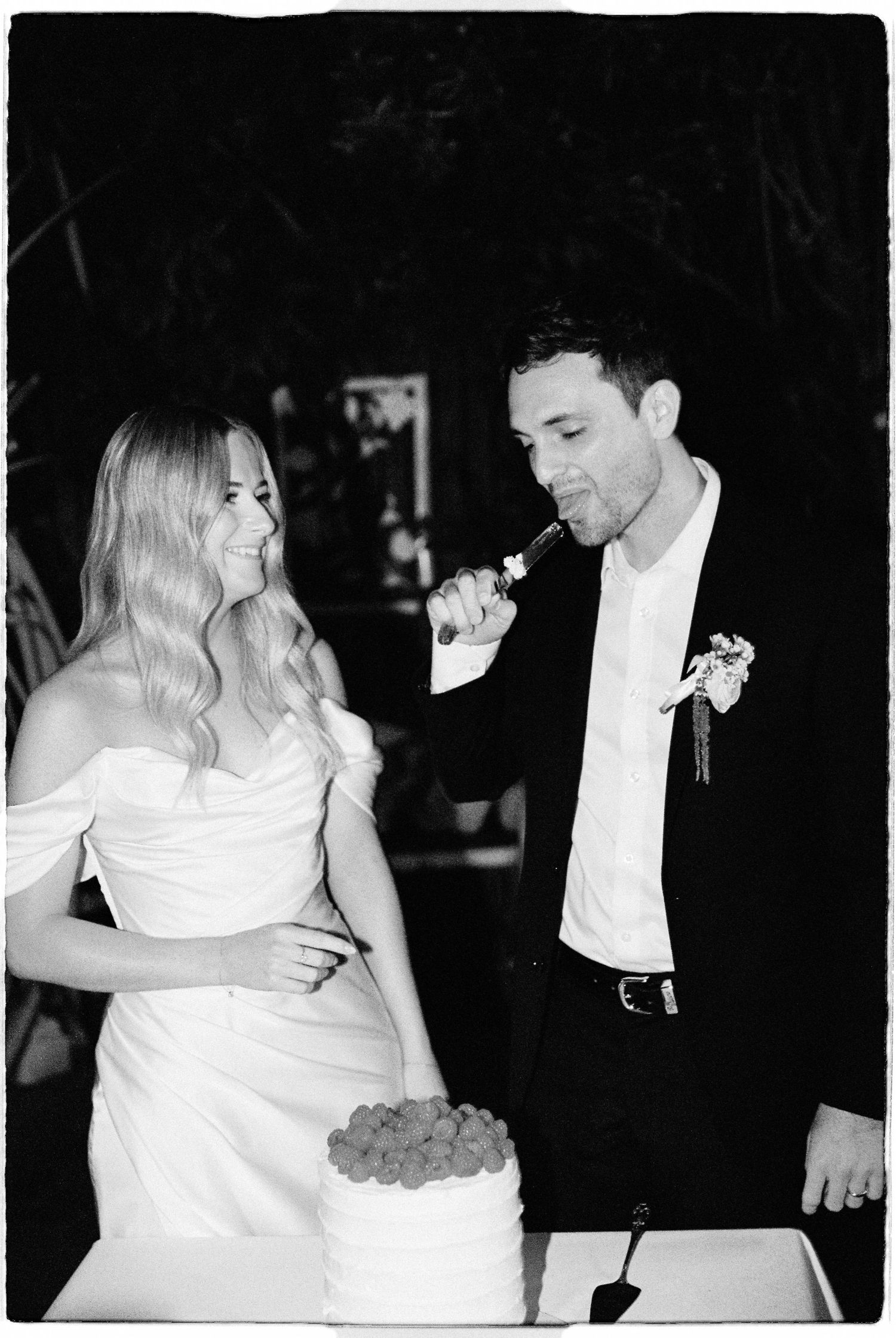Bride smiling as groom tastes wedding cake at outdoor evening reception in black and white.