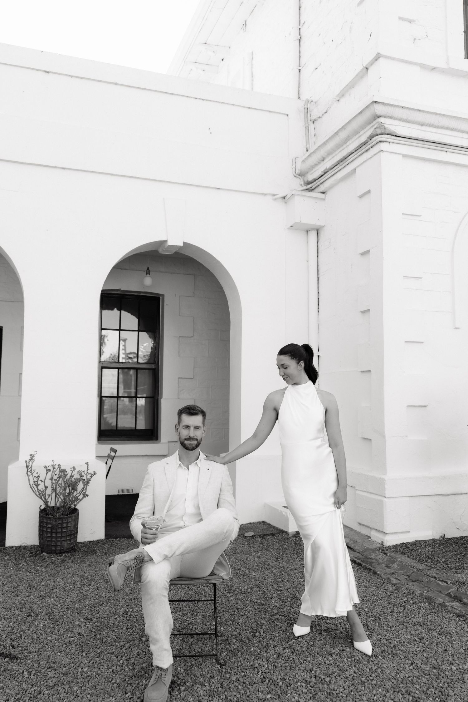 Black and white photo of a couple in white wedding attire posing elegantly outside a historic white building.