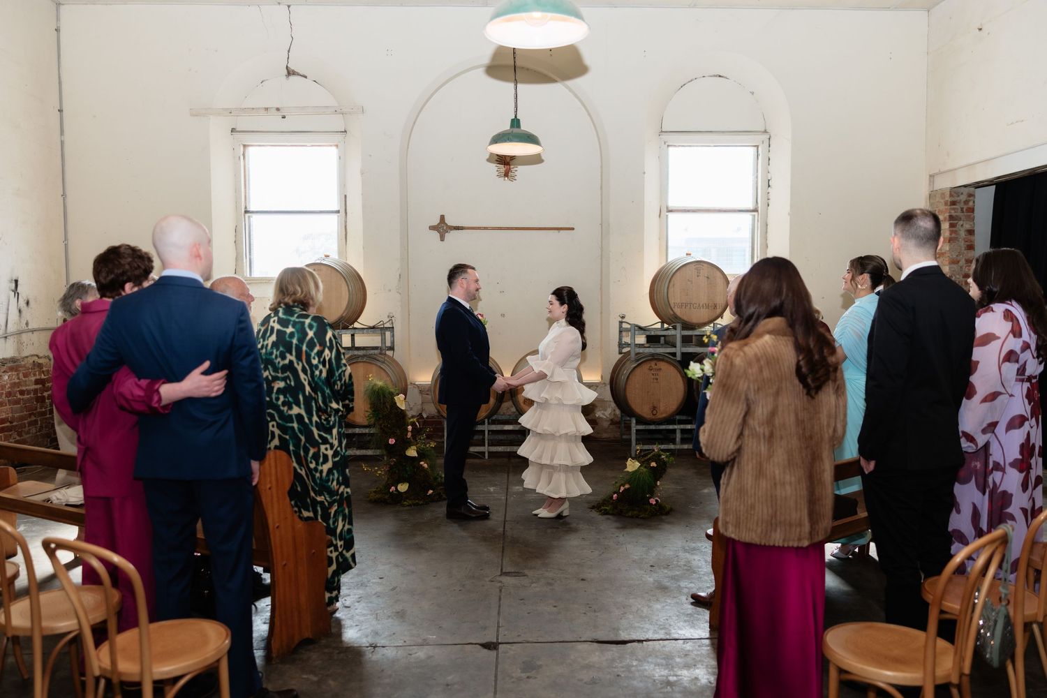 Intimate wedding ceremony in a rustic winery with wooden barrels, guests standing in rows, bride and groom facing each other.