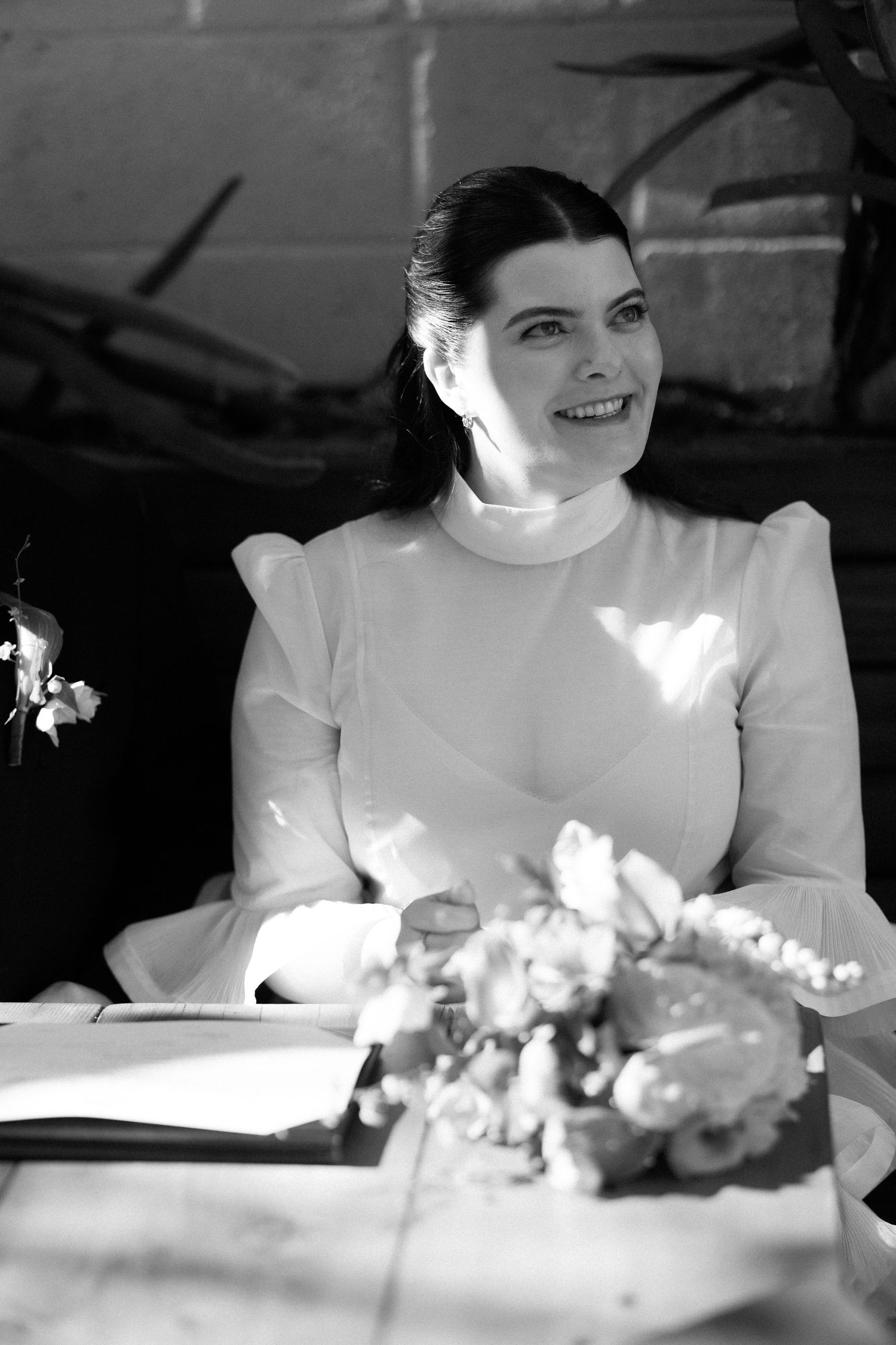 Black and white portrait of a radiant bride in a white high-neck dress smiling at her wedding signing table.