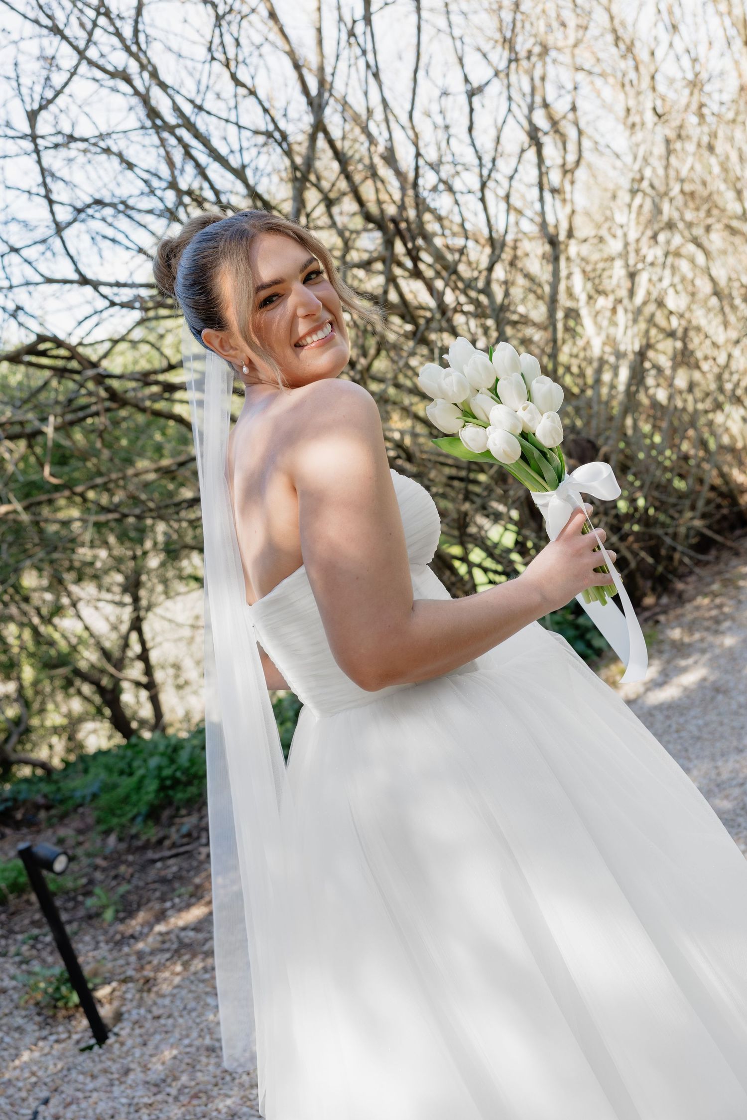 Smiling bride in white strapless gown holding white tulip bouquet, posing outdoors with veil flowing behind her.
