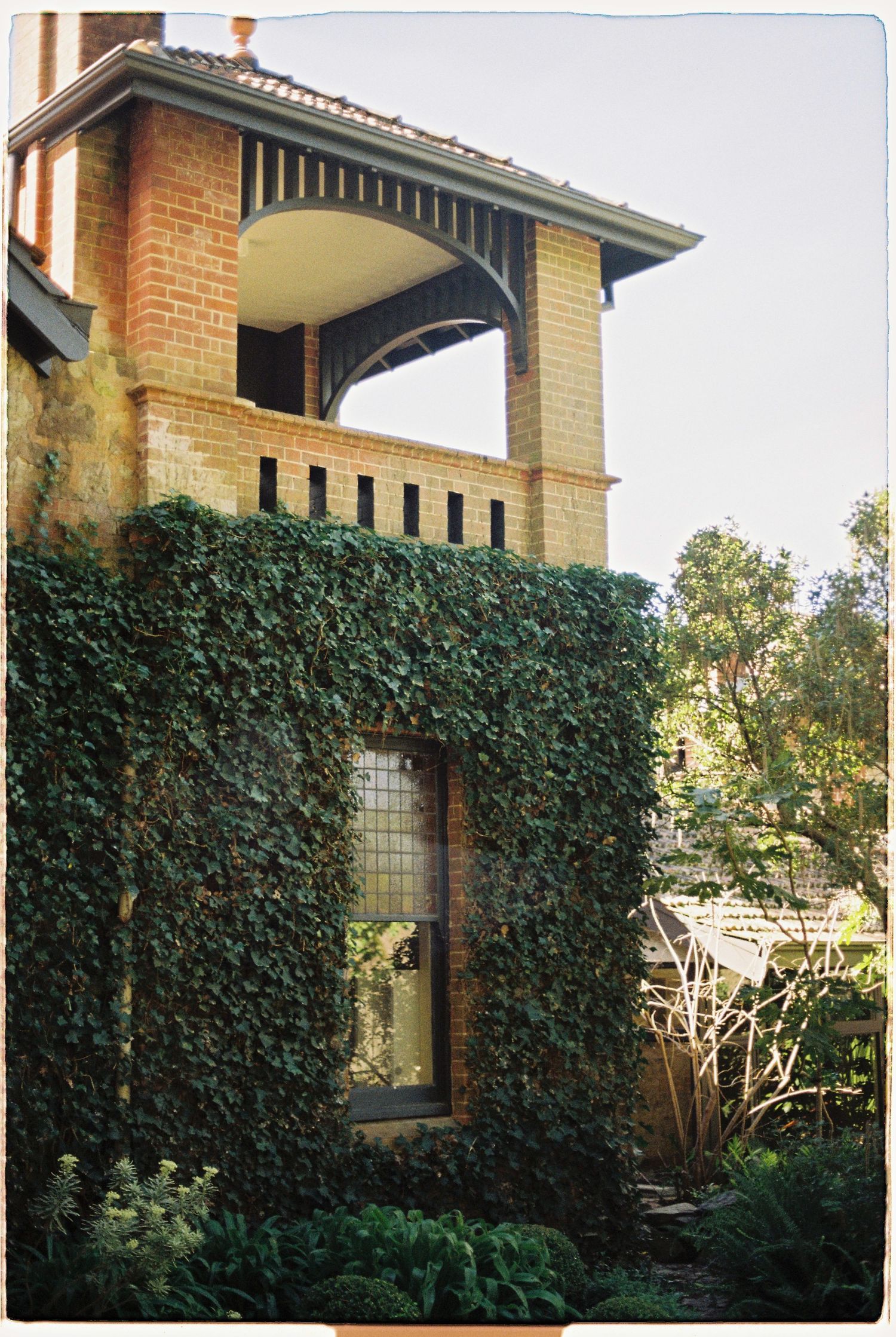 Ivy-covered brick building with arched balcony, leaded glass window, and lush garden surroundings.