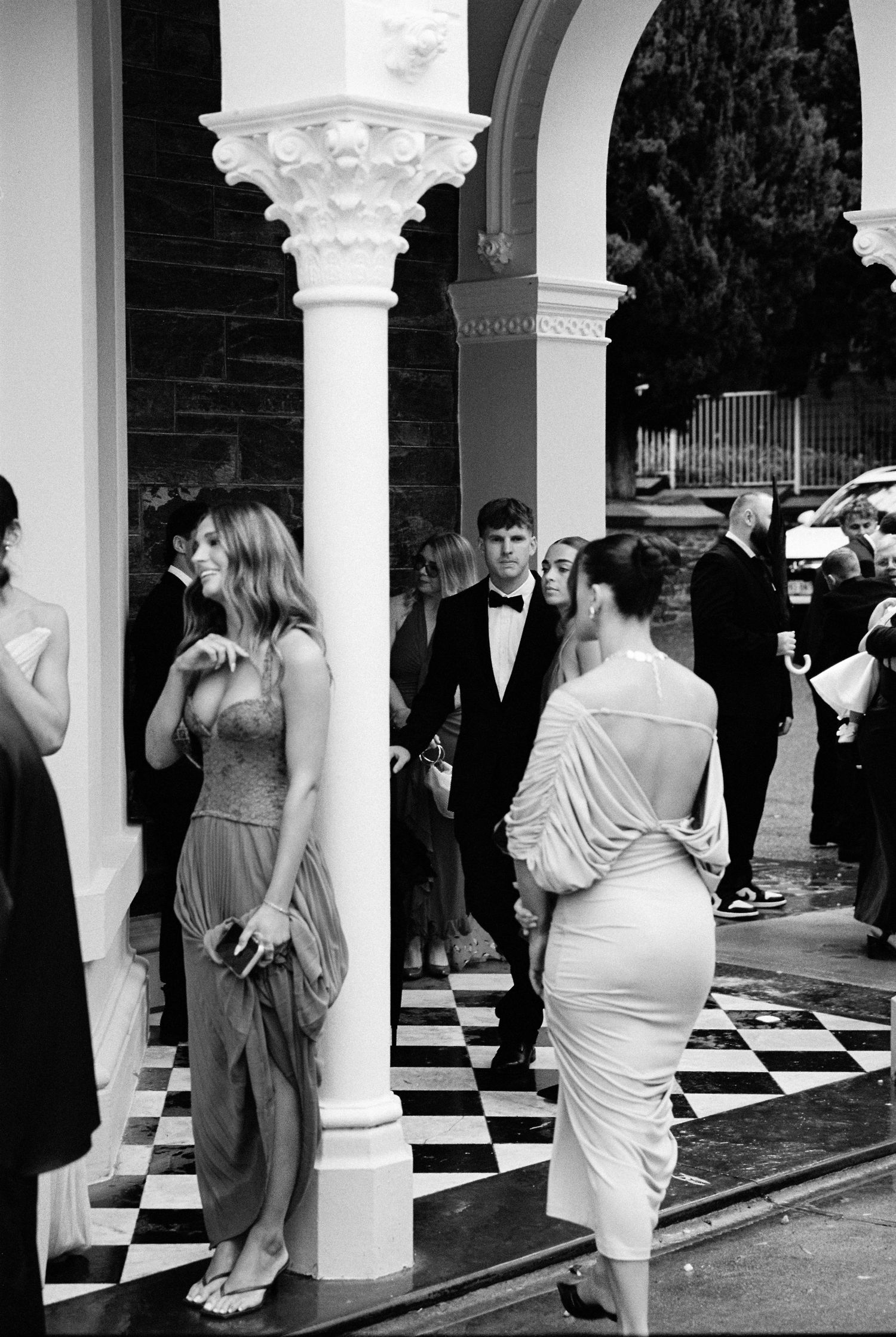 Black and white photo of elegantly dressed guests at a formal event on a checkered floor beside ornate columns.