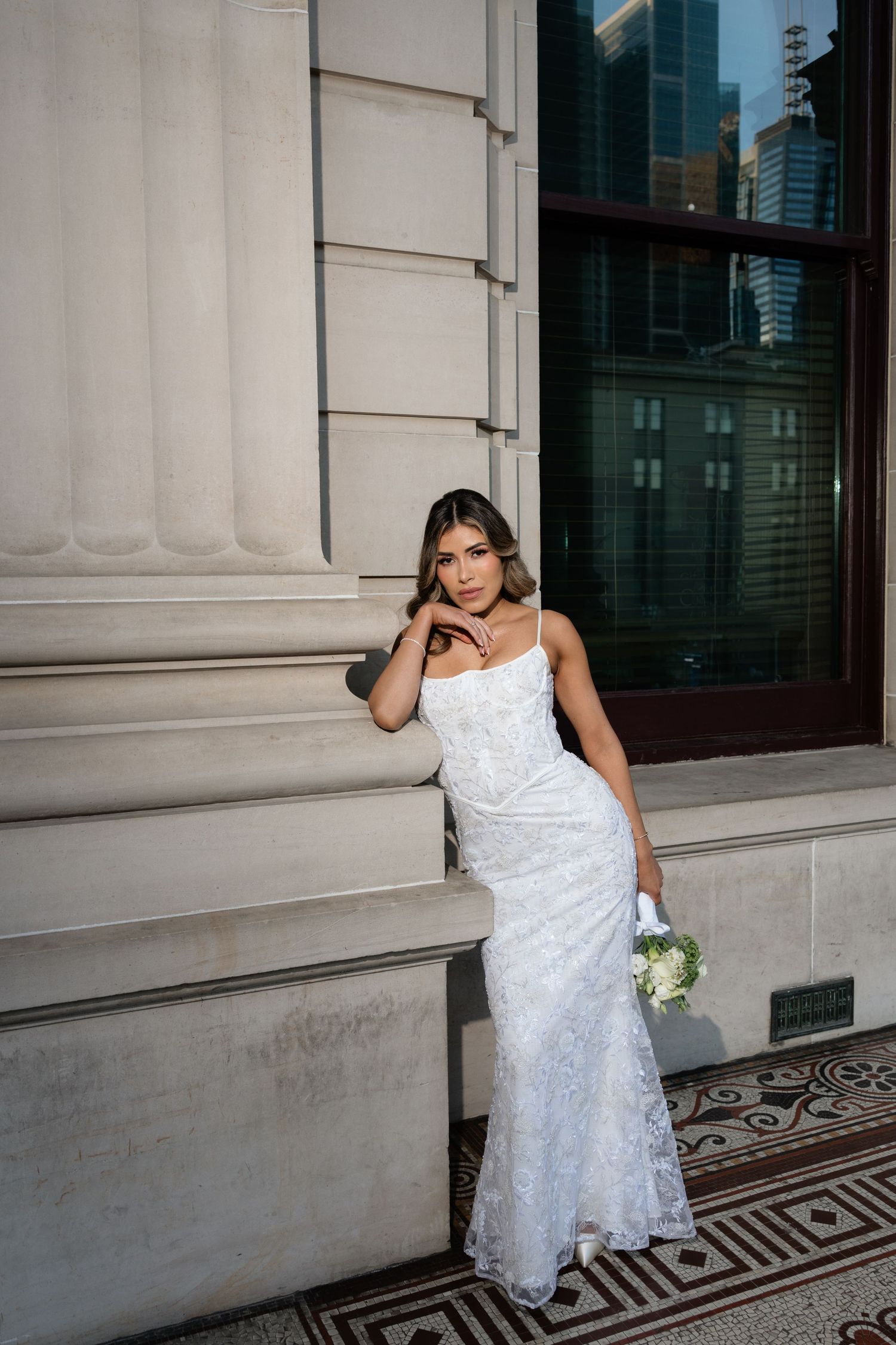Bride in white lace gown holding bouquet leaning against stone column of elegant building.