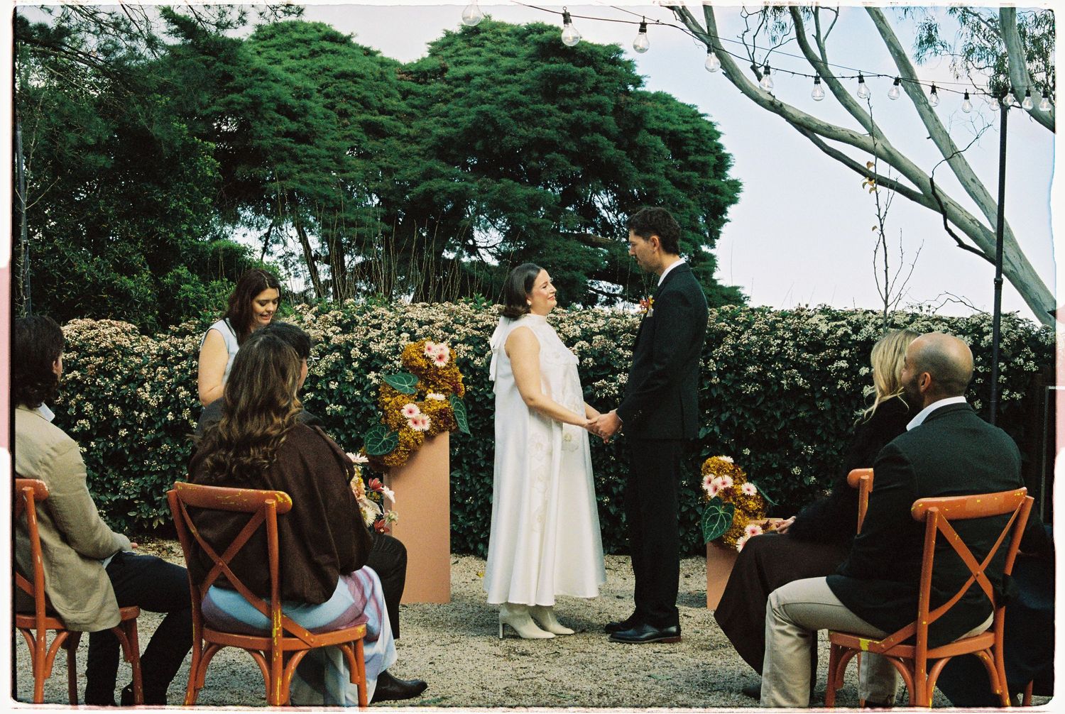 Outdoor wedding ceremony with couple exchanging vows before guests seated on wooden chairs, surrounded by lush greenery.