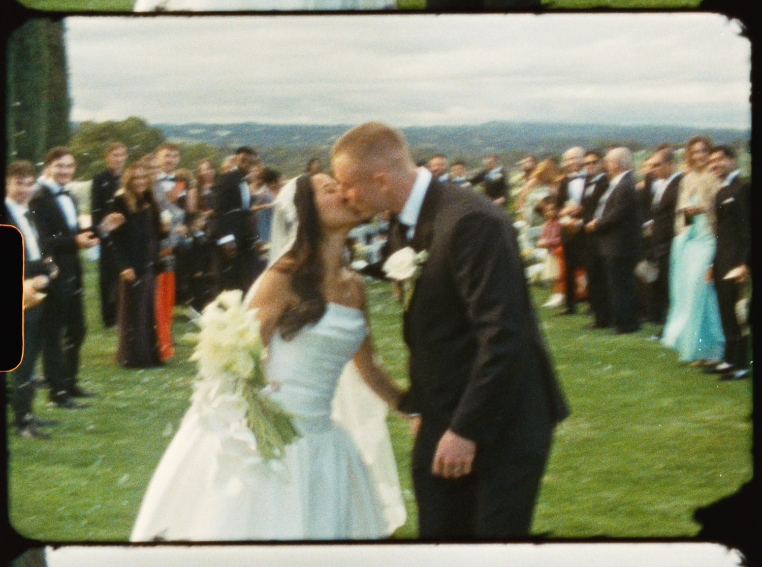 Newlyweds sharing first kiss as married couple while wedding guests celebrate around them outdoors.