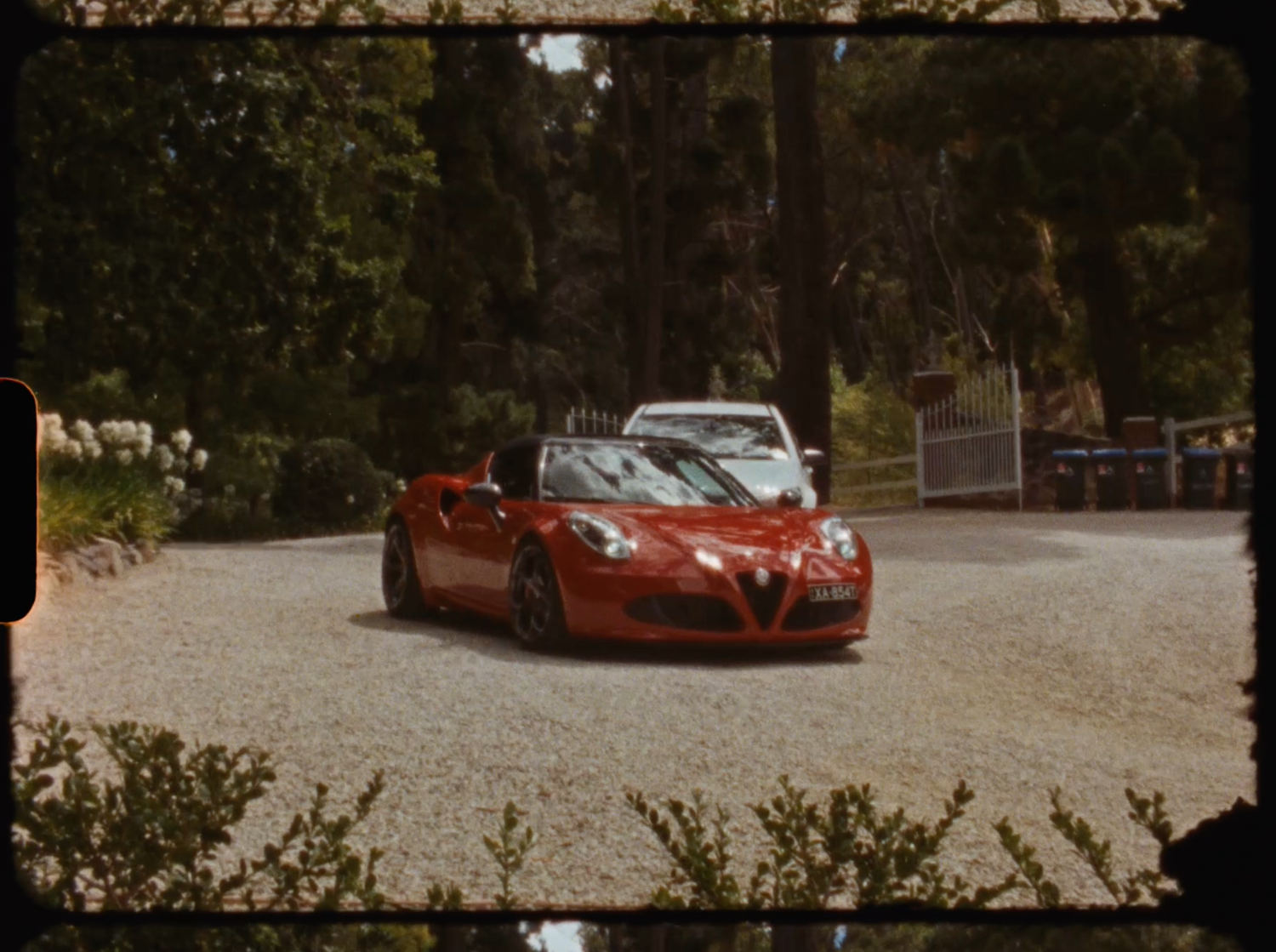 Red Alfa Romeo sports car driving on a gravel path surrounded by trees.
