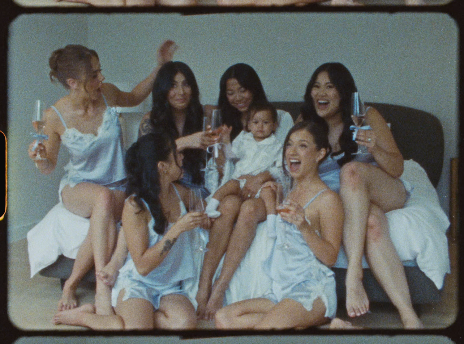 Group of women in blue satin pajamas celebrating on a bed with champagne glasses and a baby.