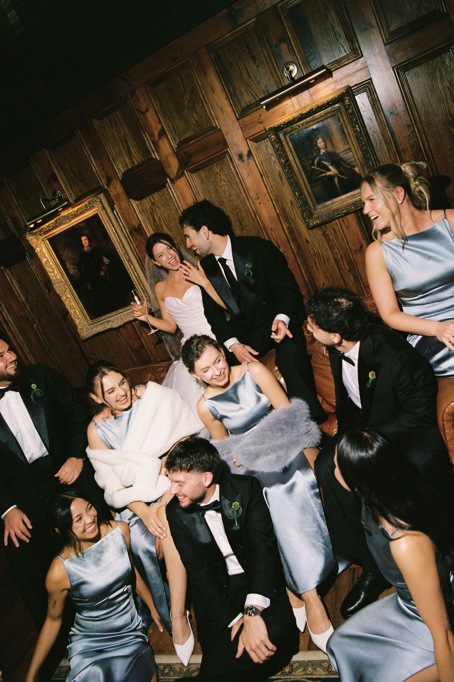 Wedding party laughing and celebrating together in a wood-paneled room with oil paintings.