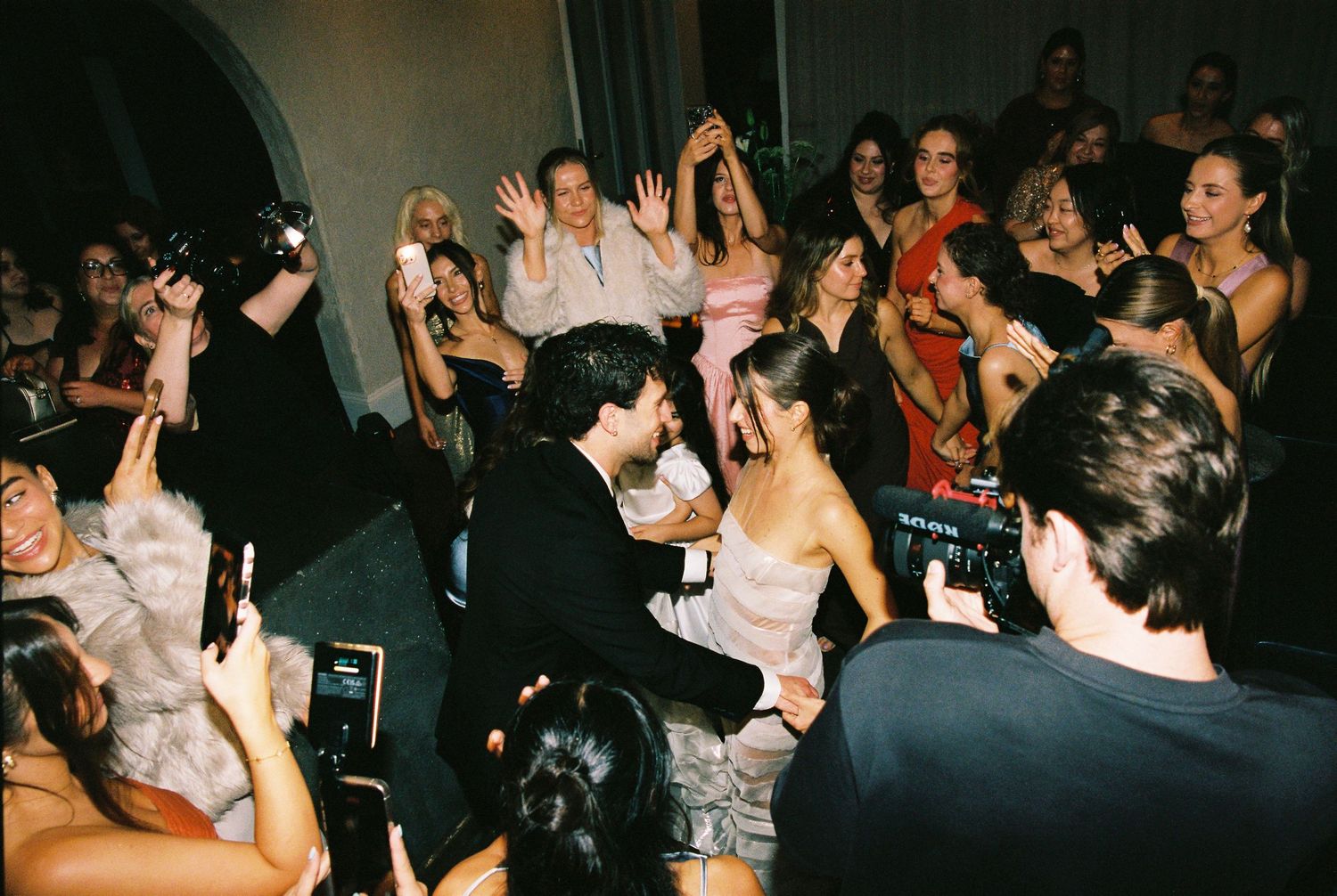 Bride and groom share first dance surrounded by cheering wedding guests capturing the moment on phones.