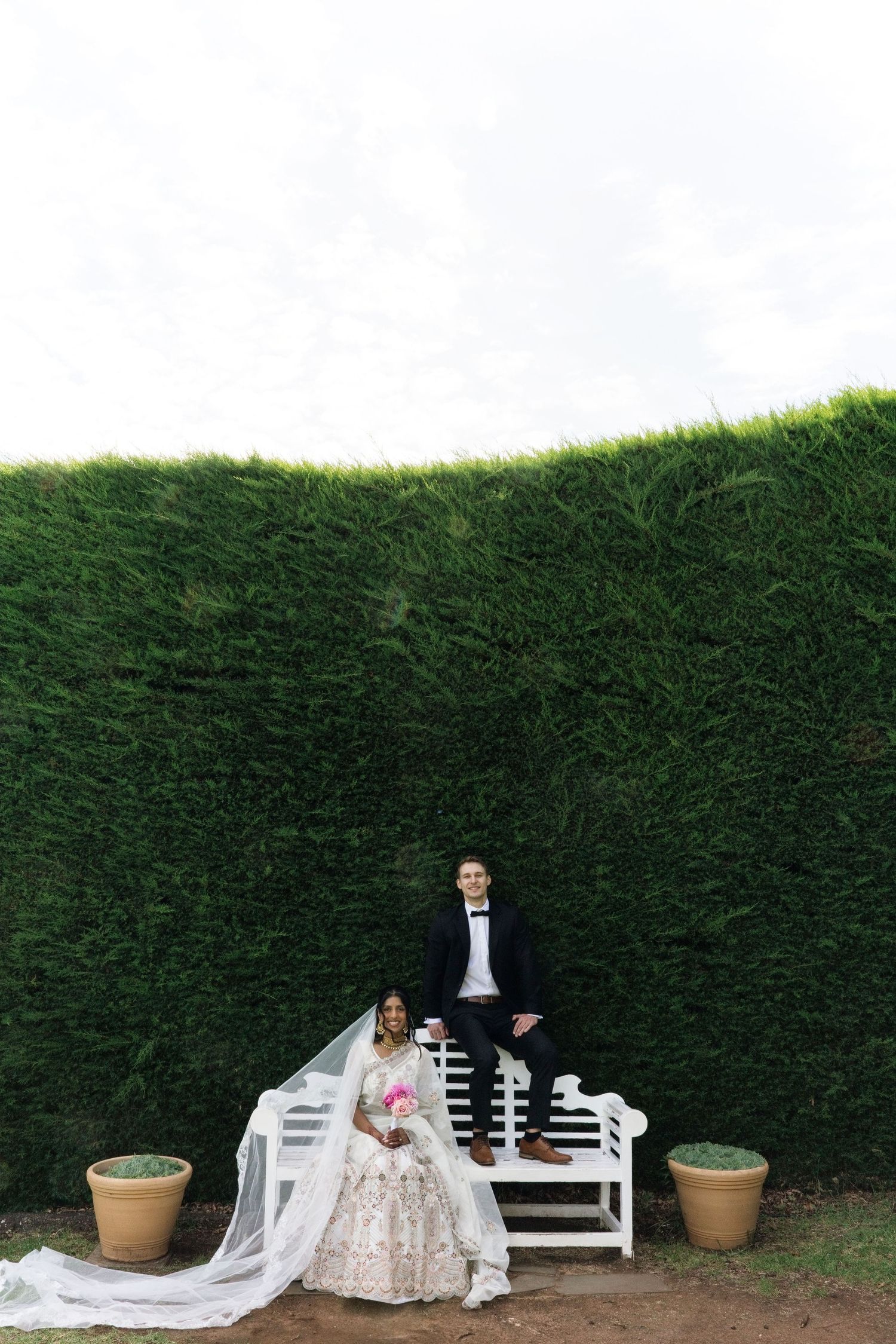 Bride in white lehenga with long veil seated on bench, groom standing behind, tall green hedge backdrop.