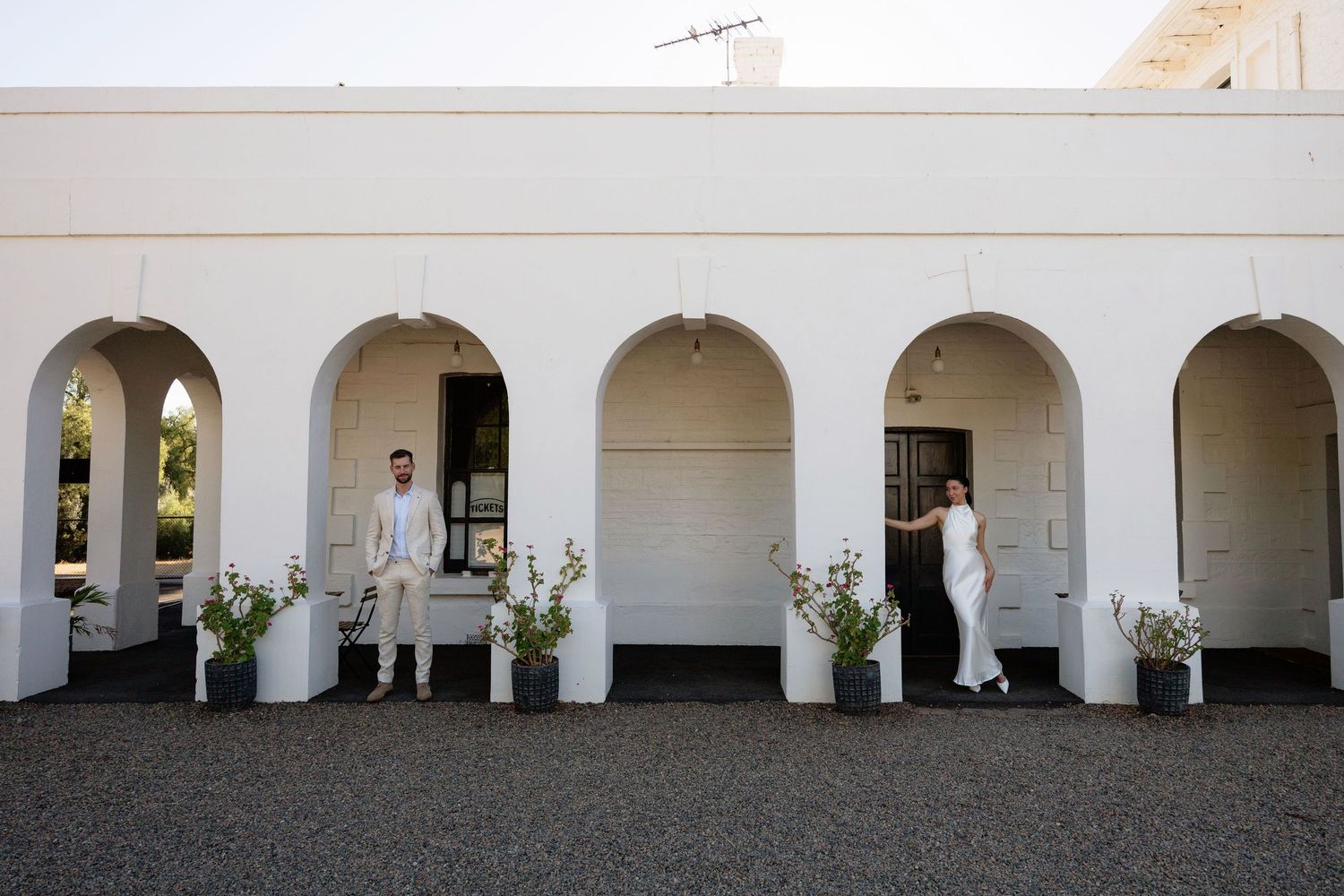 Bride and groom stand apart under white arched colonnade of historic wedding venue.