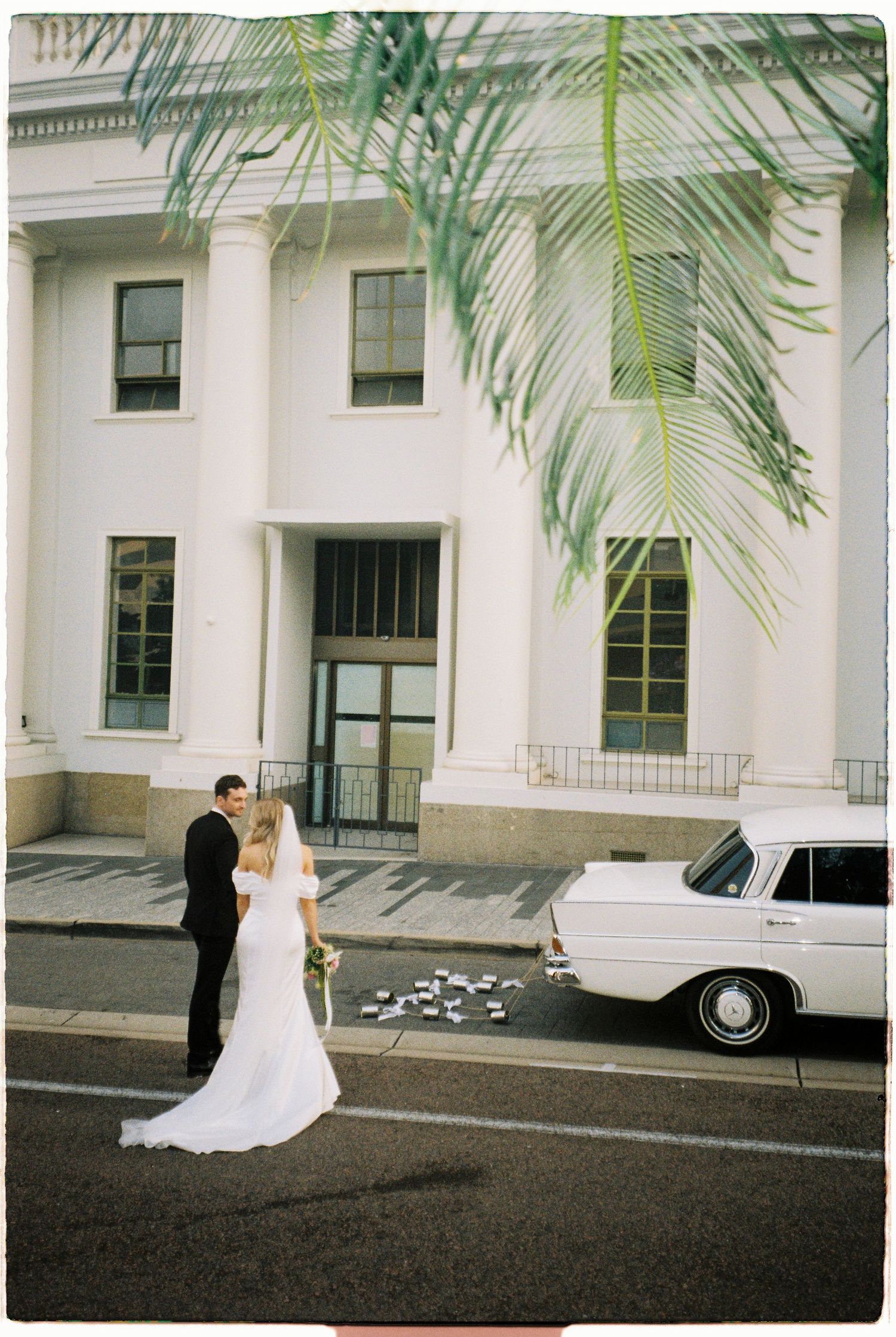 Elegant bride and groom embracing outside grand white colonial building beside vintage wedding car.