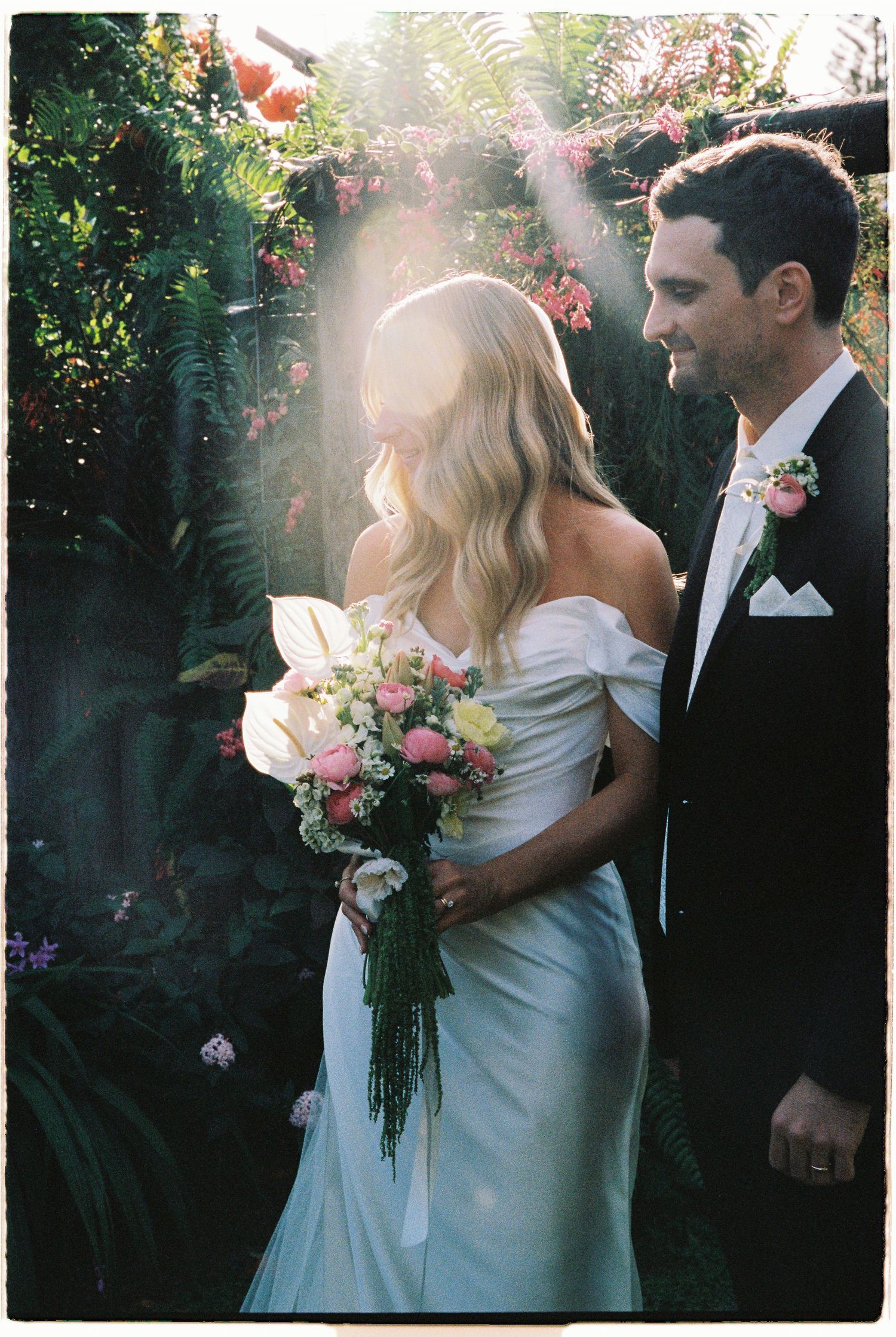 Bride holding pink bouquet in off-shoulder gown with groom in tuxedo in sunlit garden setting.