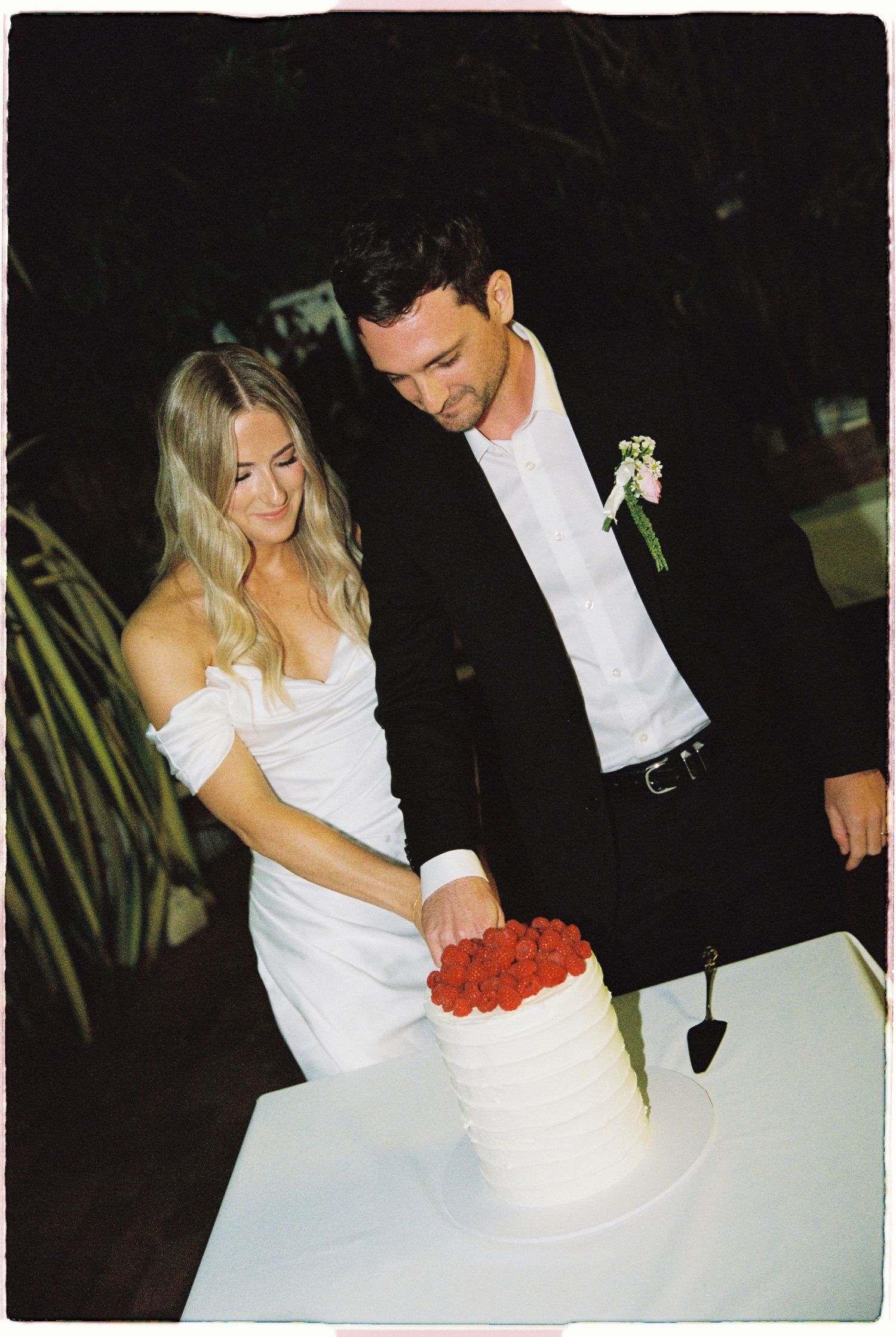 Bride and groom cut white wedding cake topped with fresh red berries at outdoor evening reception.