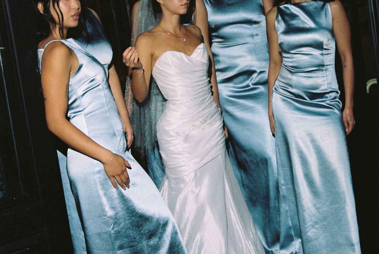 Bride in white gown with bridesmaids in light blue satin dresses posing together at a wedding.