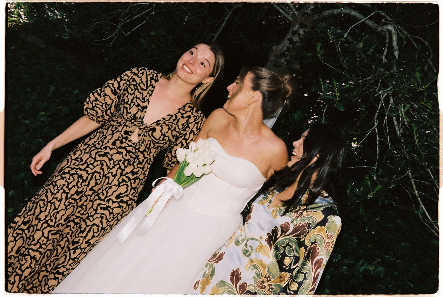 Bride in white wedding dress with bouquet laughing with two guests in floral dresses outdoors.