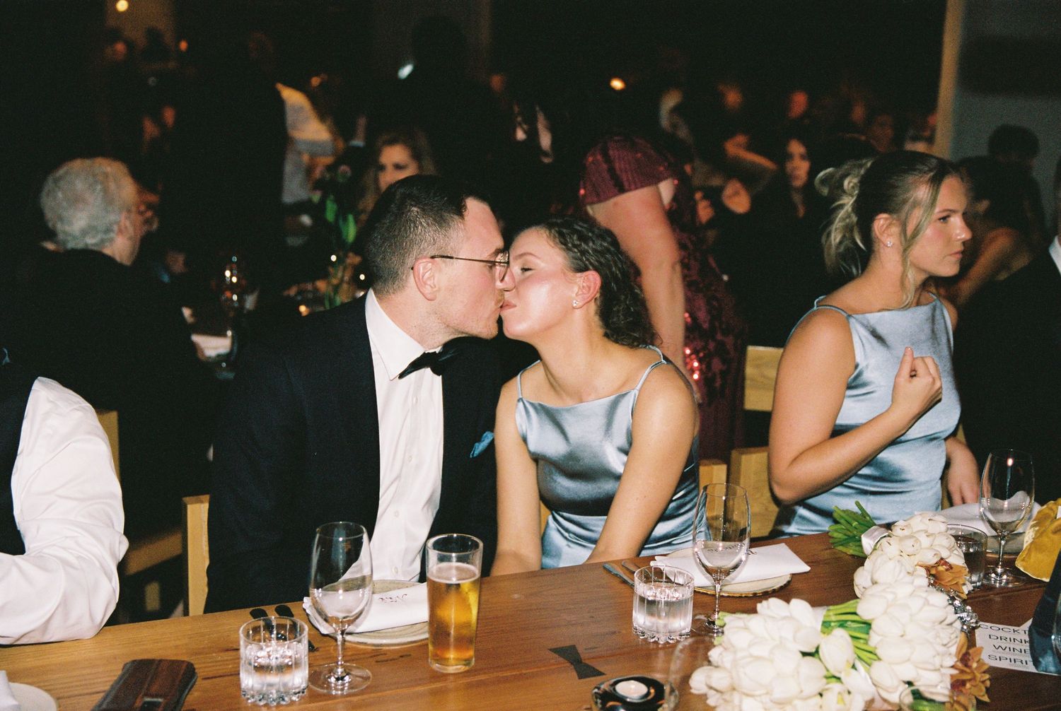 Couple shares a kiss at wedding reception table with beer glasses and flowers.